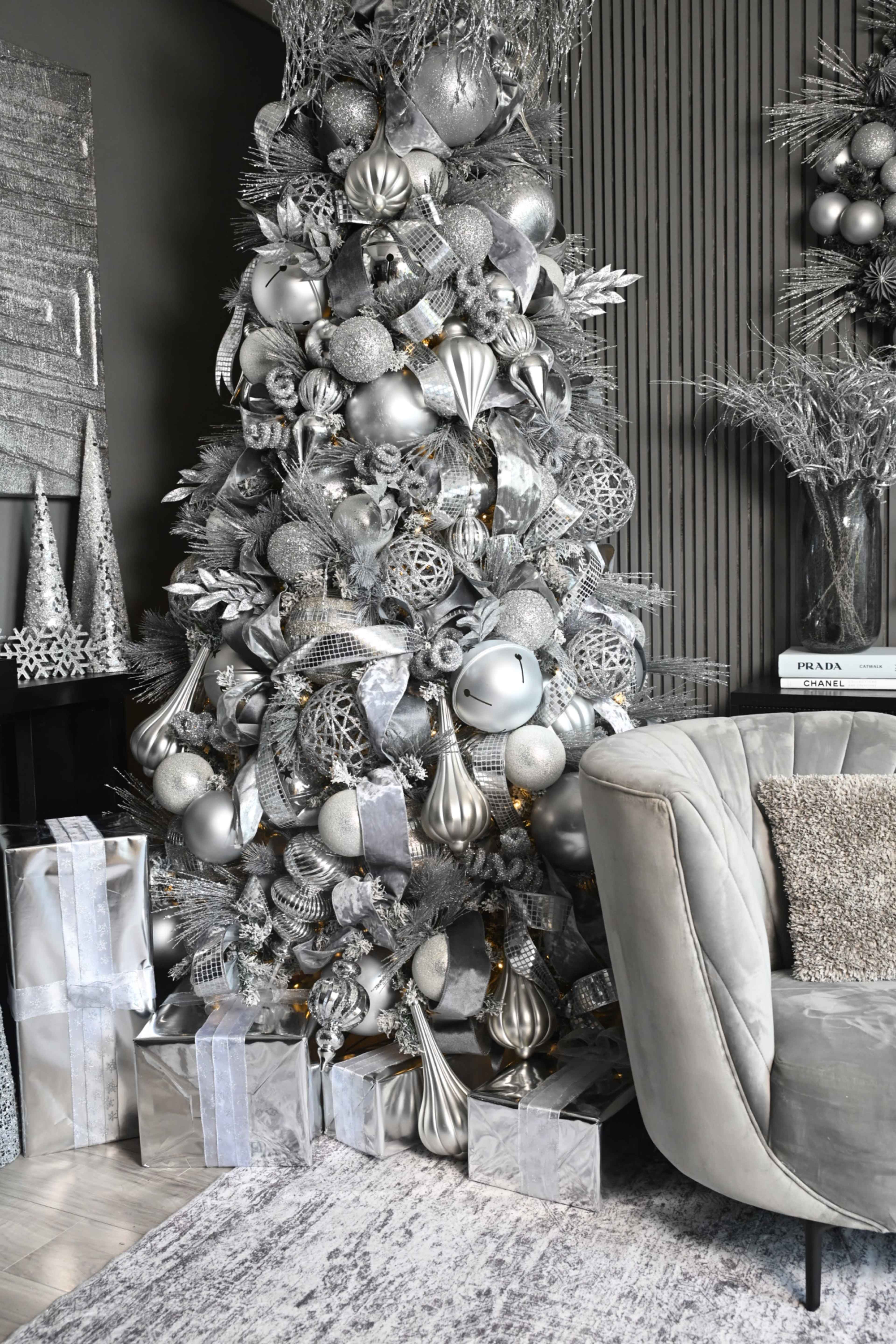A decorated silver Christmas tree is positioned next to a plush gray sofa, surrounded by wrapped gifts.