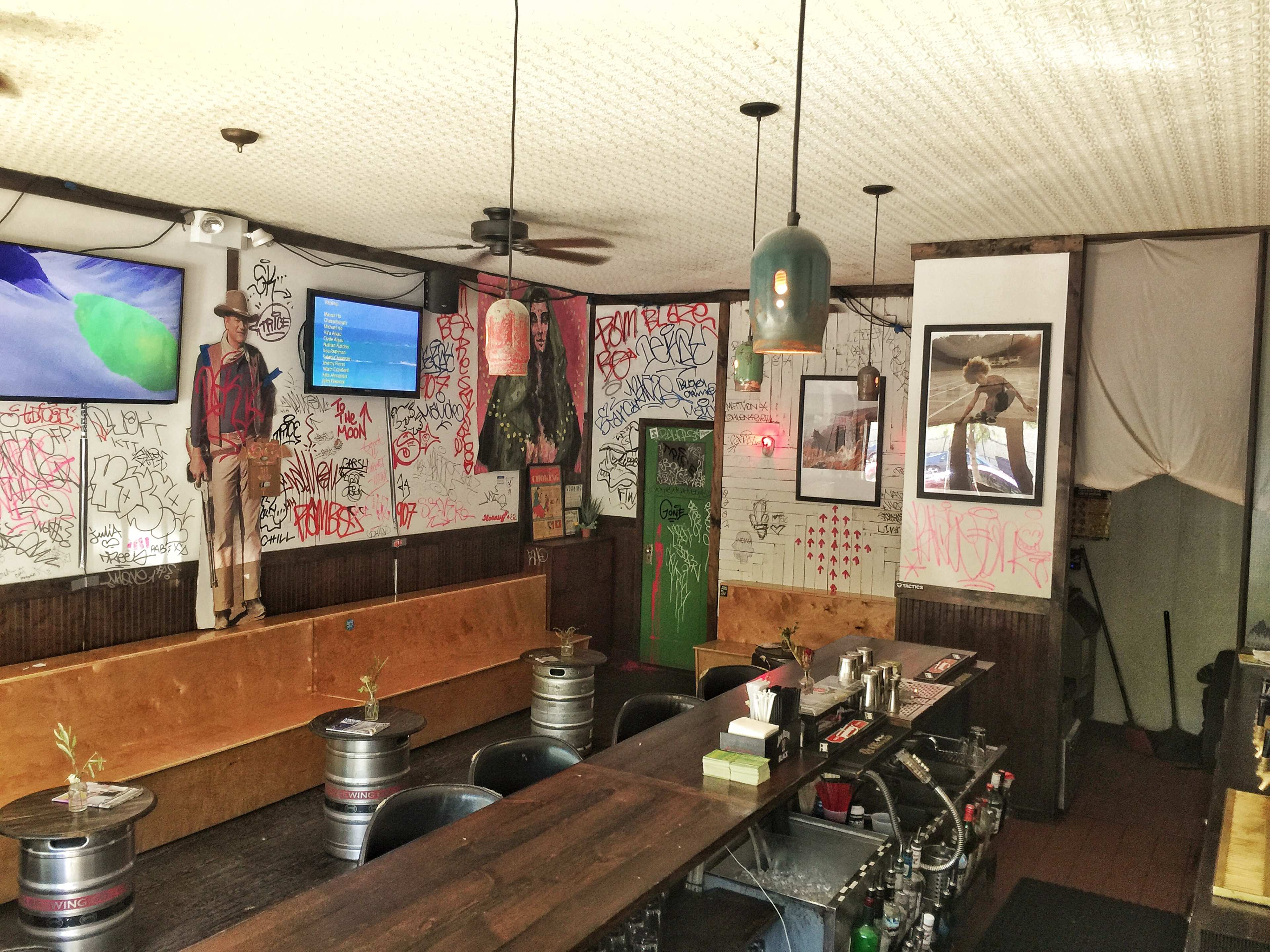 The interior of a bar featuring graffiti-covered walls, a long wooden counter, and two televisions displaying content.