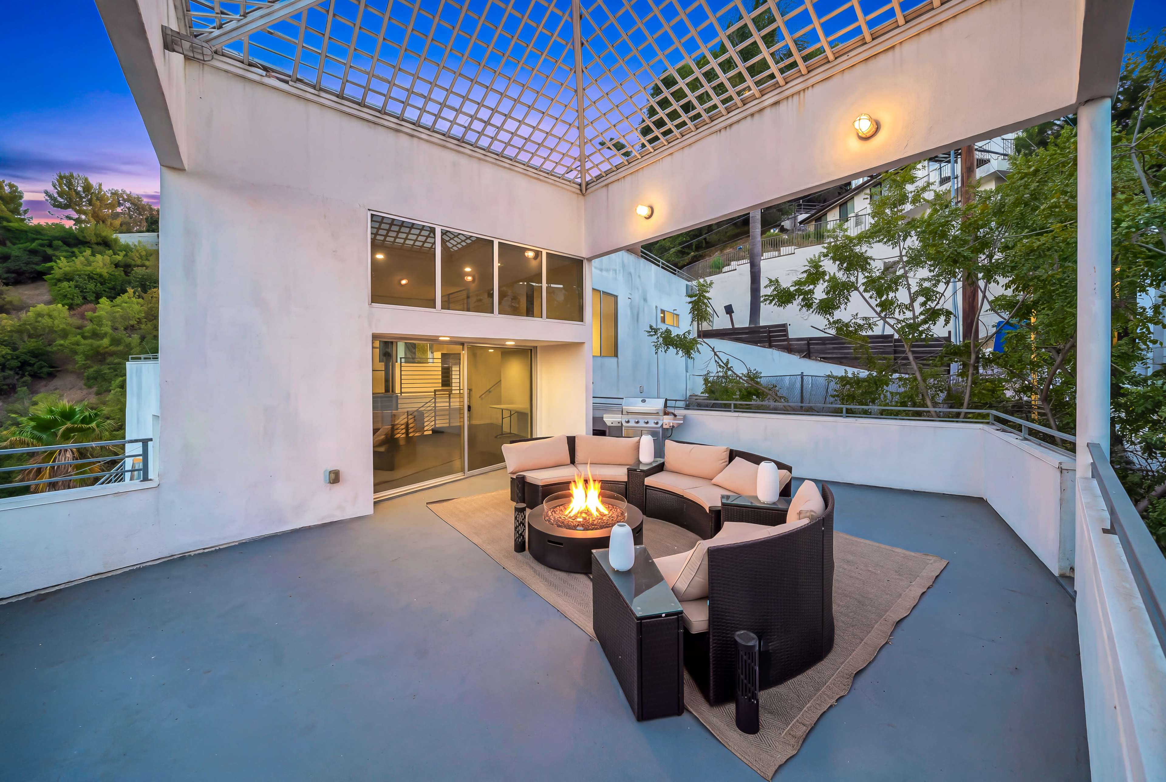 The image shows a modern outdoor lounge area with a circular seating arrangement around a fire pit, set on a patio with a trellis overhead.