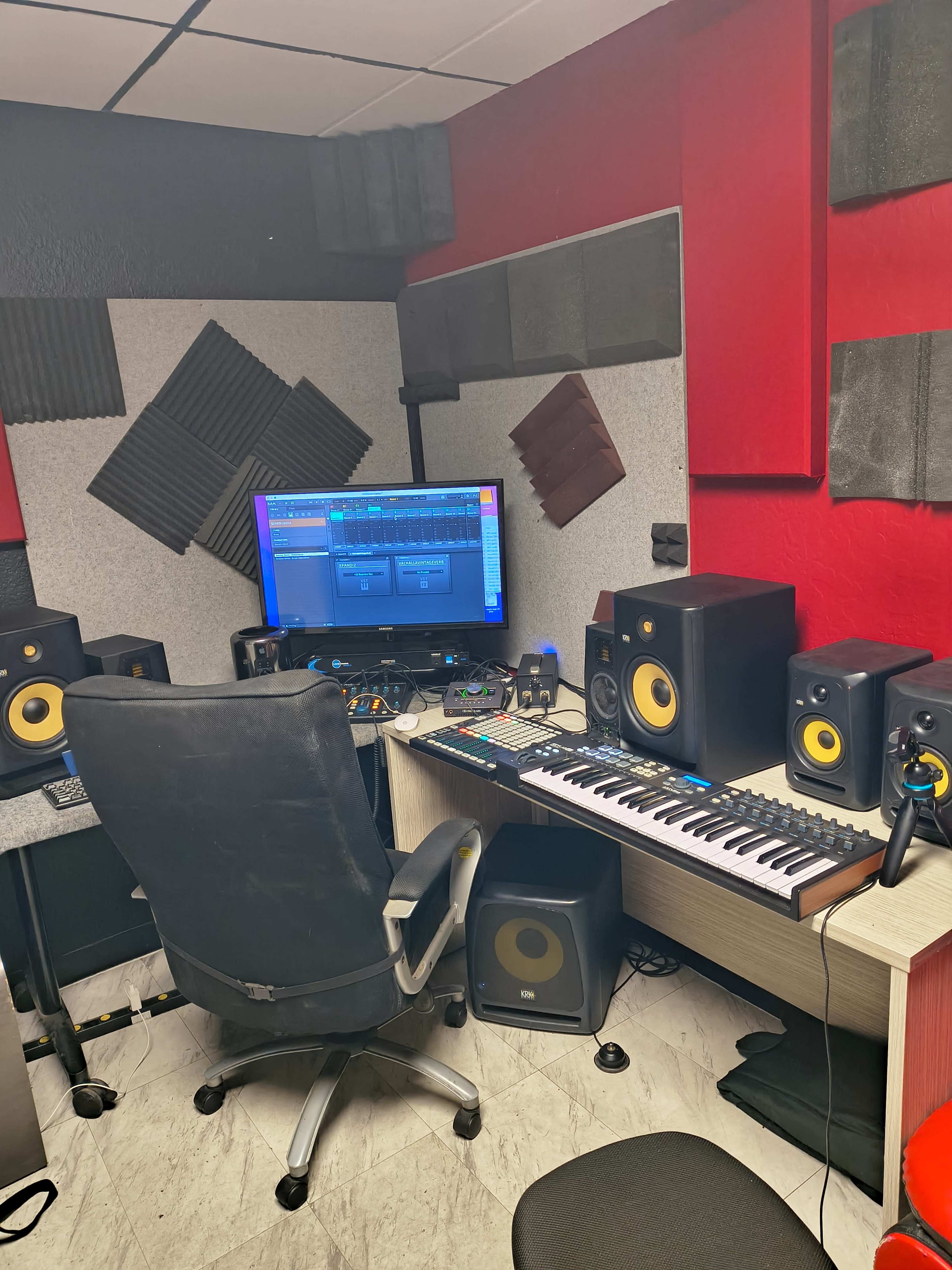 A small music production studio features a computer, audio monitors, a keyboard, and acoustic panels on the walls.