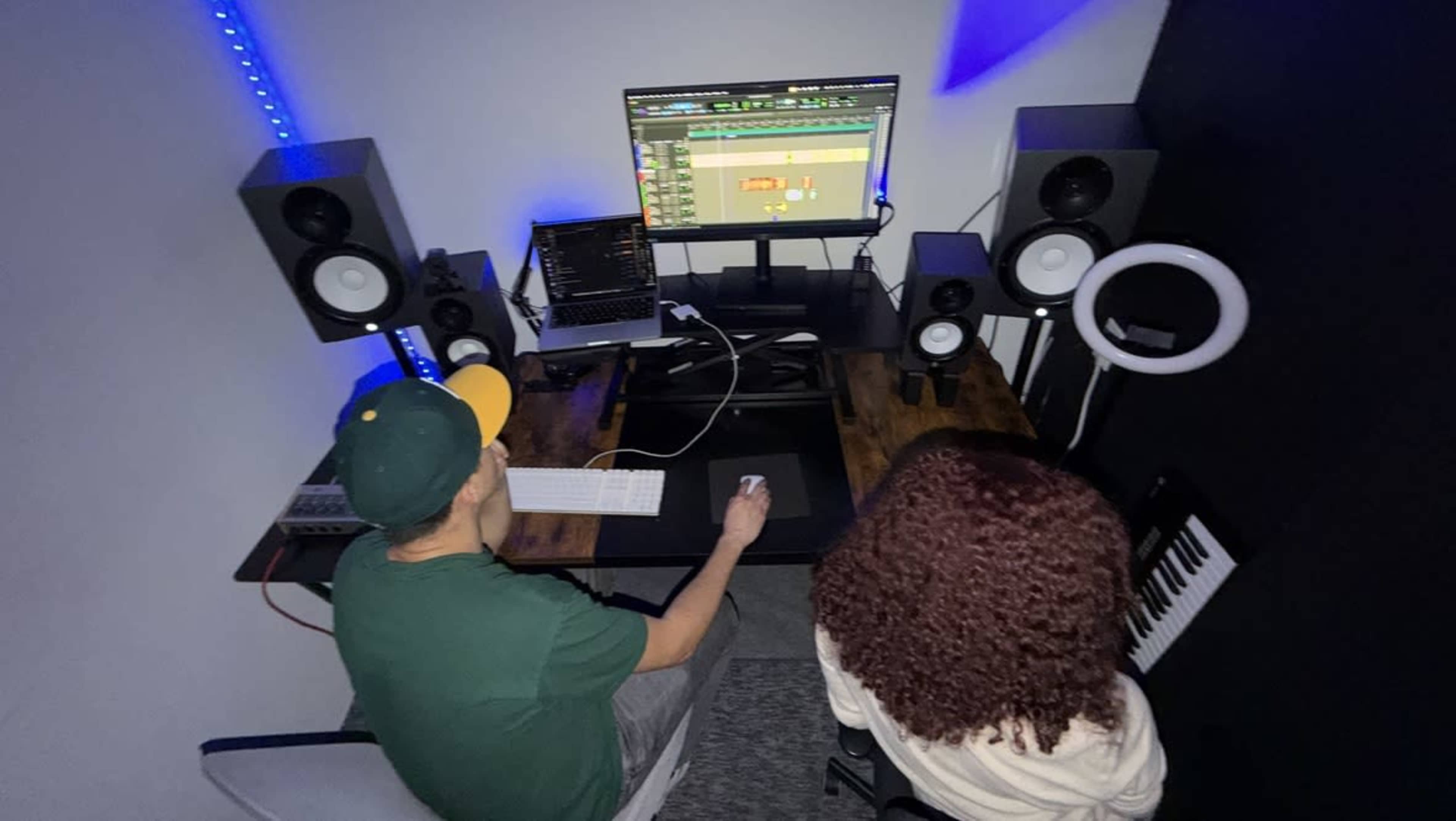 Two individuals are seated at a desk in a music production studio, one using a laptop and the other facing a computer monitor displaying audio editing software.