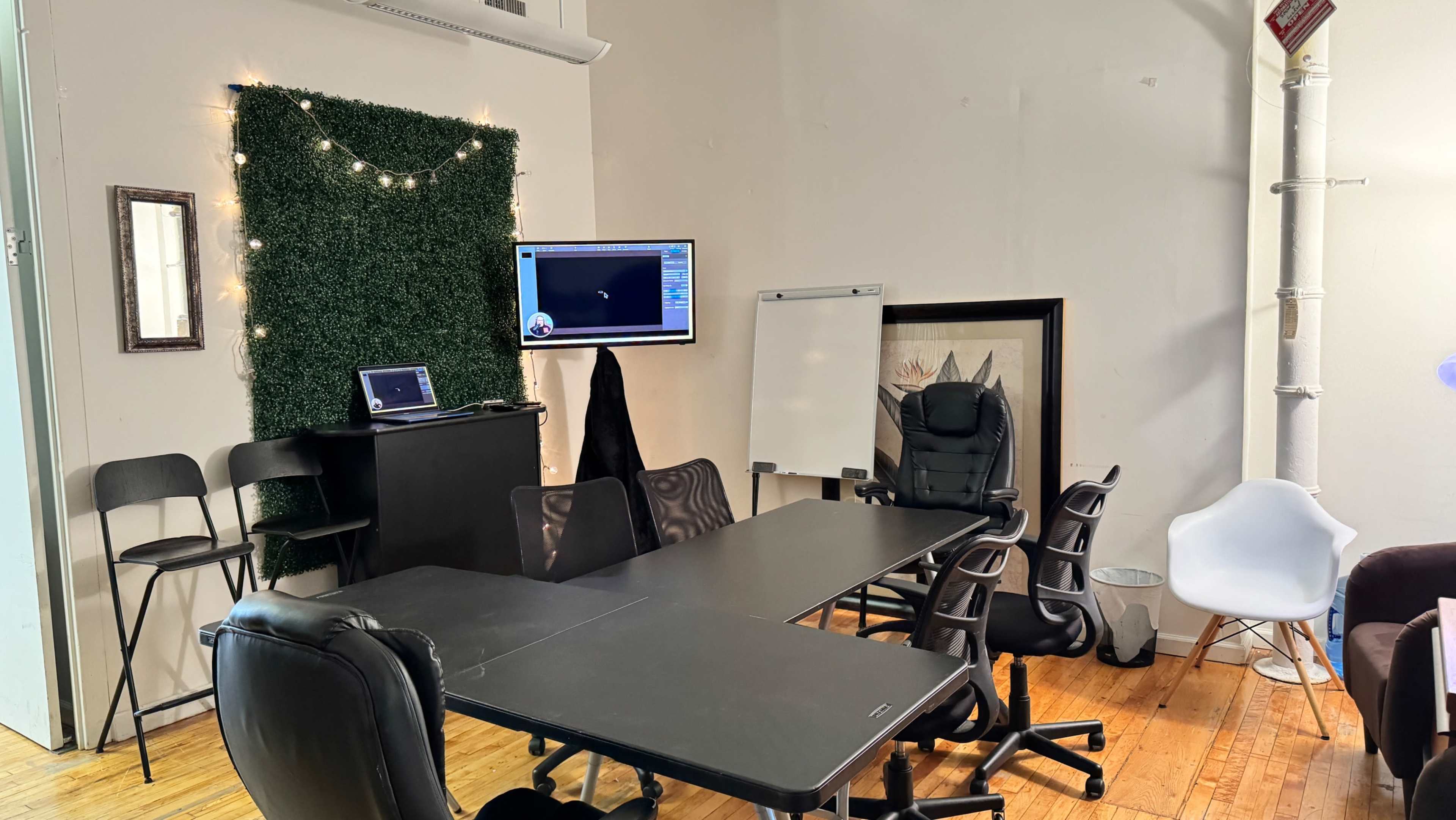 A small meeting room features several office chairs, a large table, a whiteboard, and a television mounted on the wall, with a green wall decoration in the background.