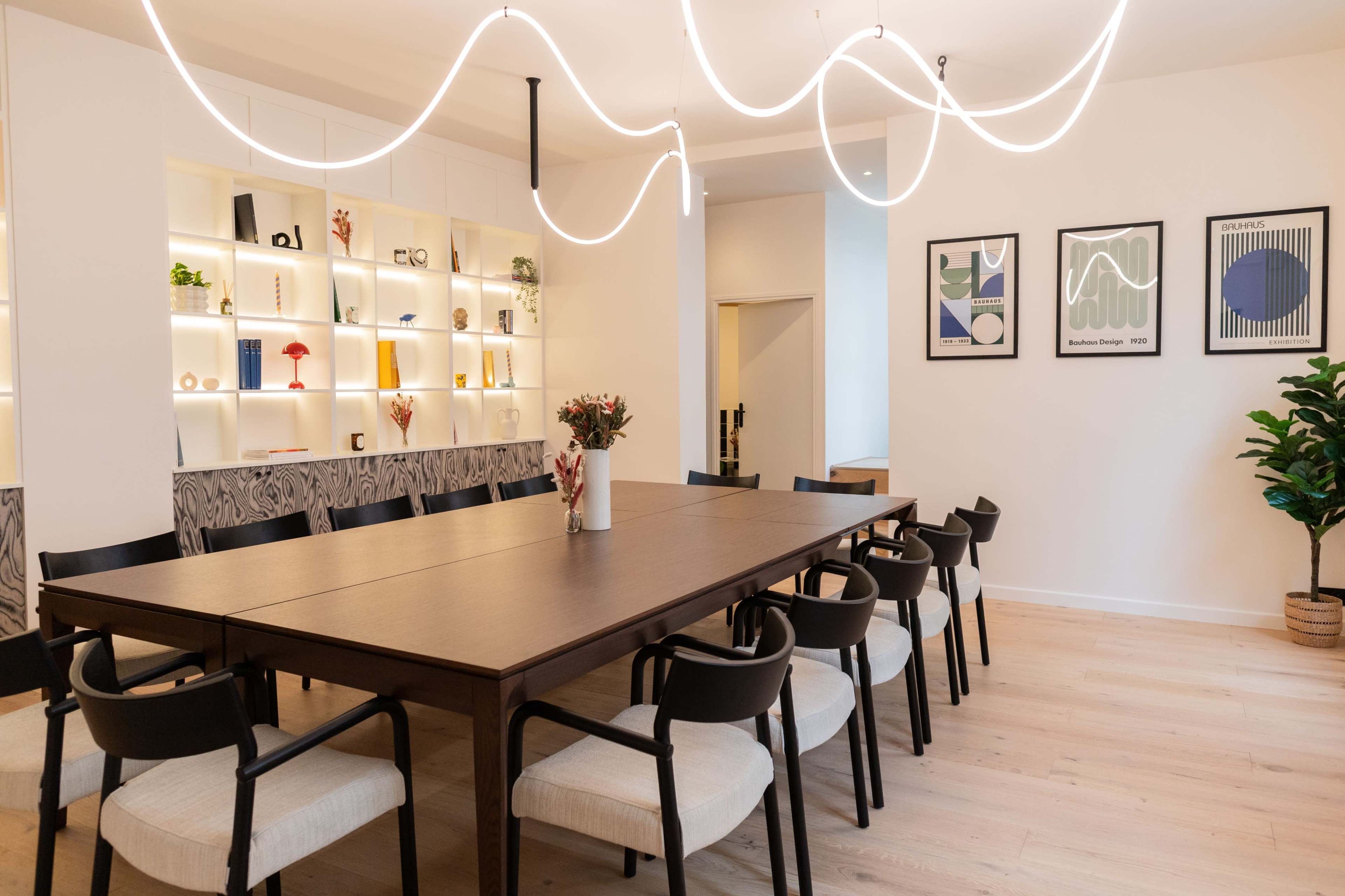 A long wooden dining table is surrounded by black and white chairs in a well-lit room with decorative shelves and framed artwork on the walls.