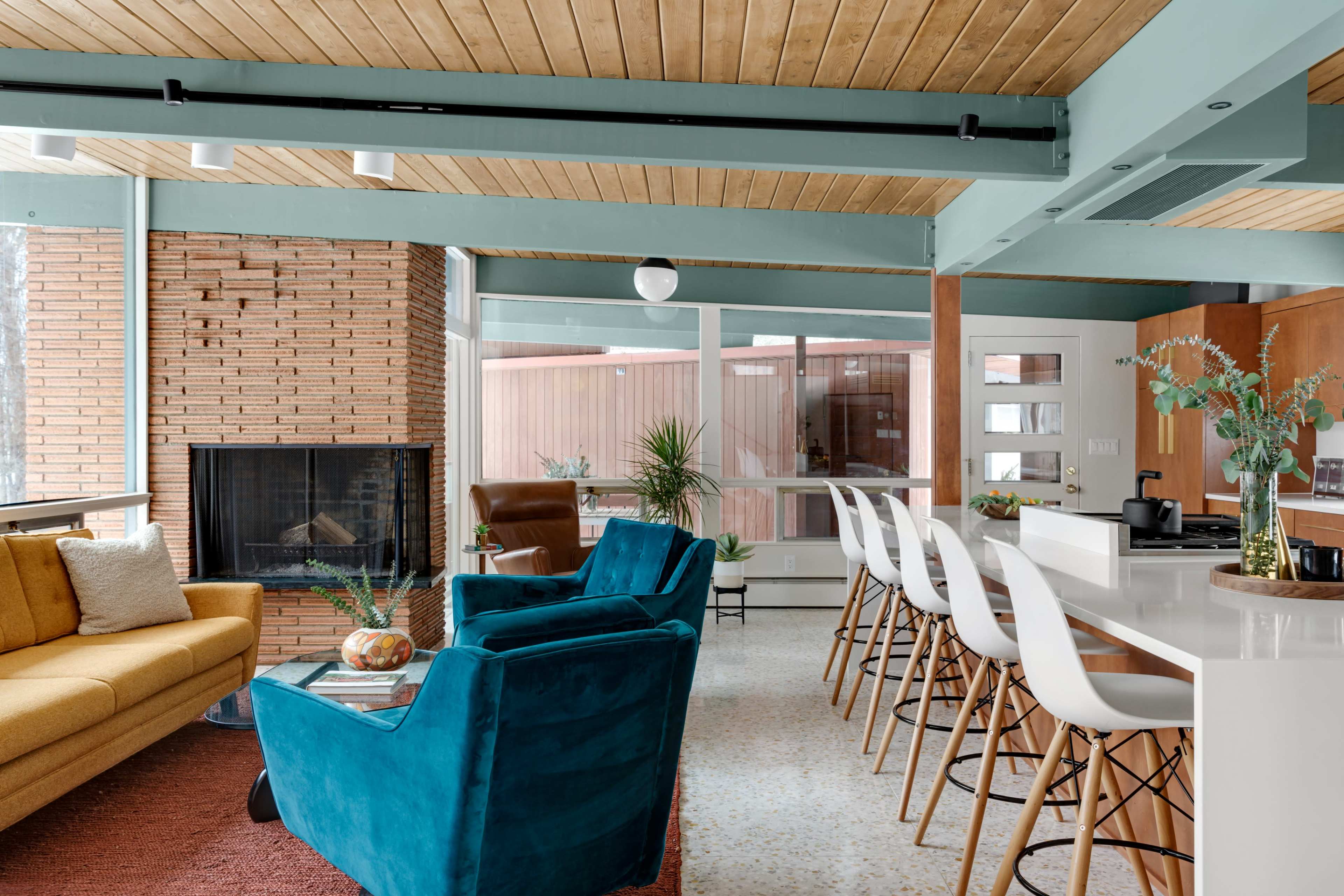 The living space features a bright yellow sofa and teal chairs surrounding a modern kitchen with bar stools and a large window offering natural light.