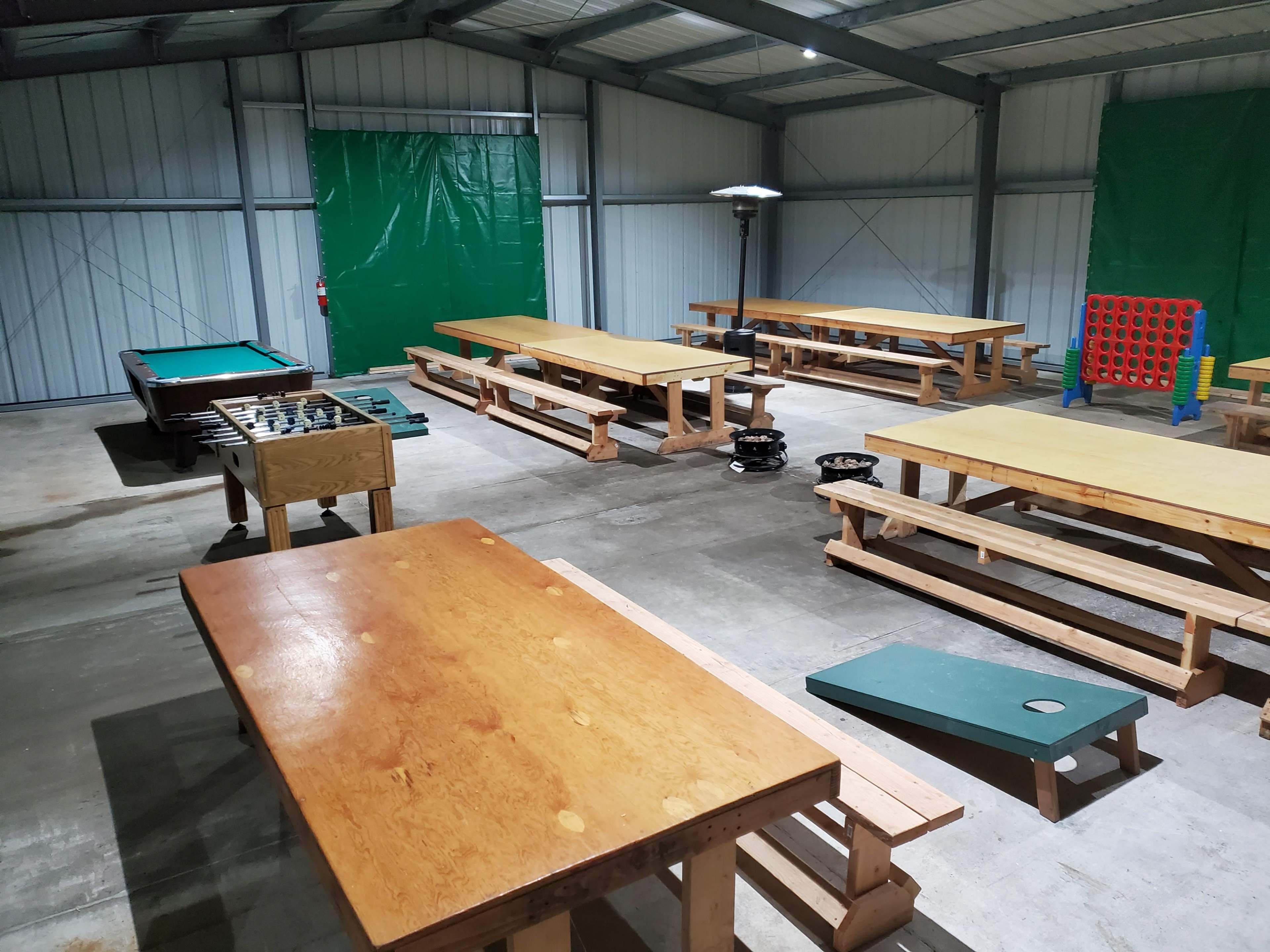 An indoor recreational space featuring several wooden tables and benches, a pool table, a foosball table, and a large Connect Four game setup.