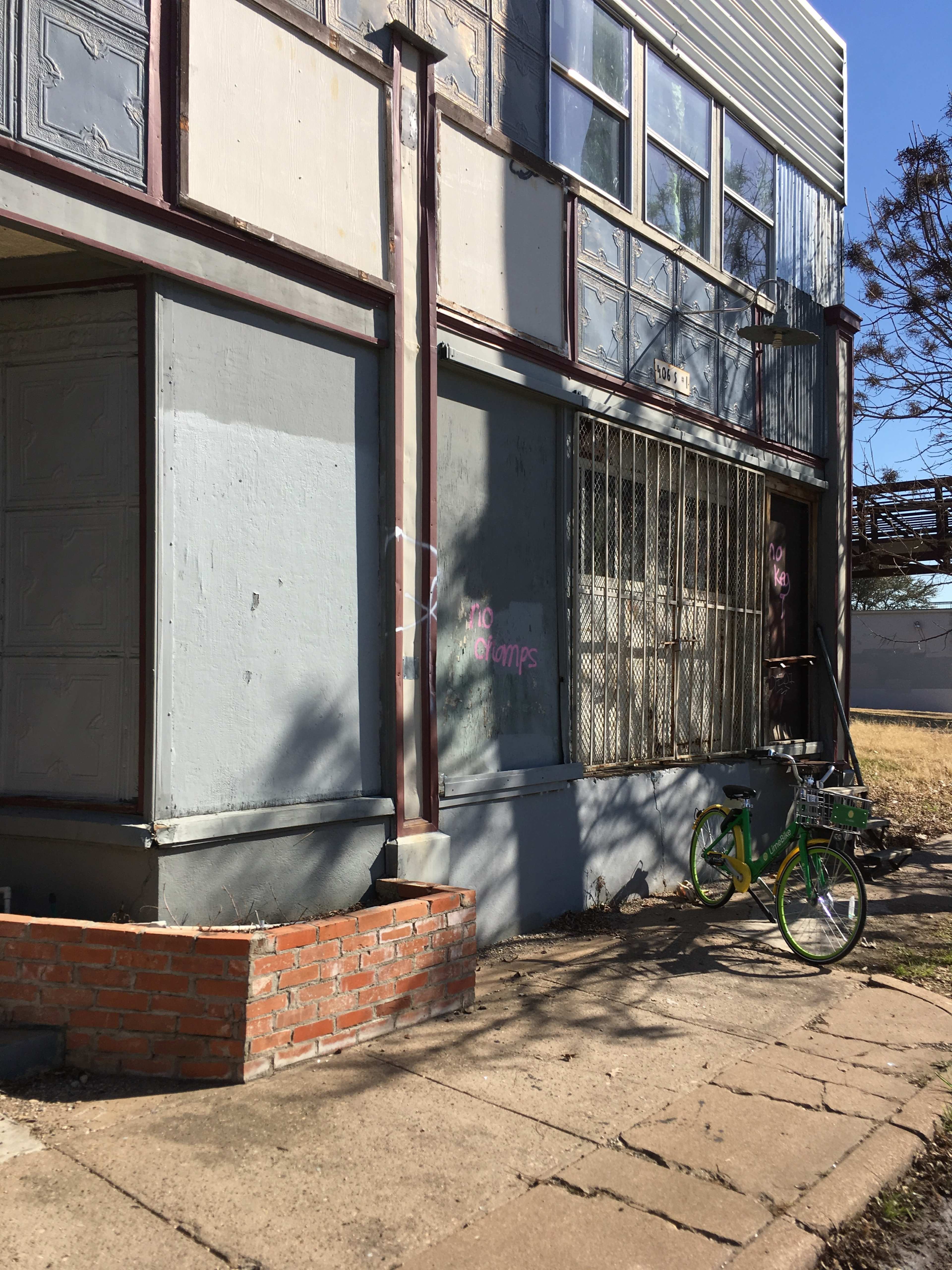 A vacant building with a concrete sidewalk features a green bicycle near the entrance and graffiti on the wall.