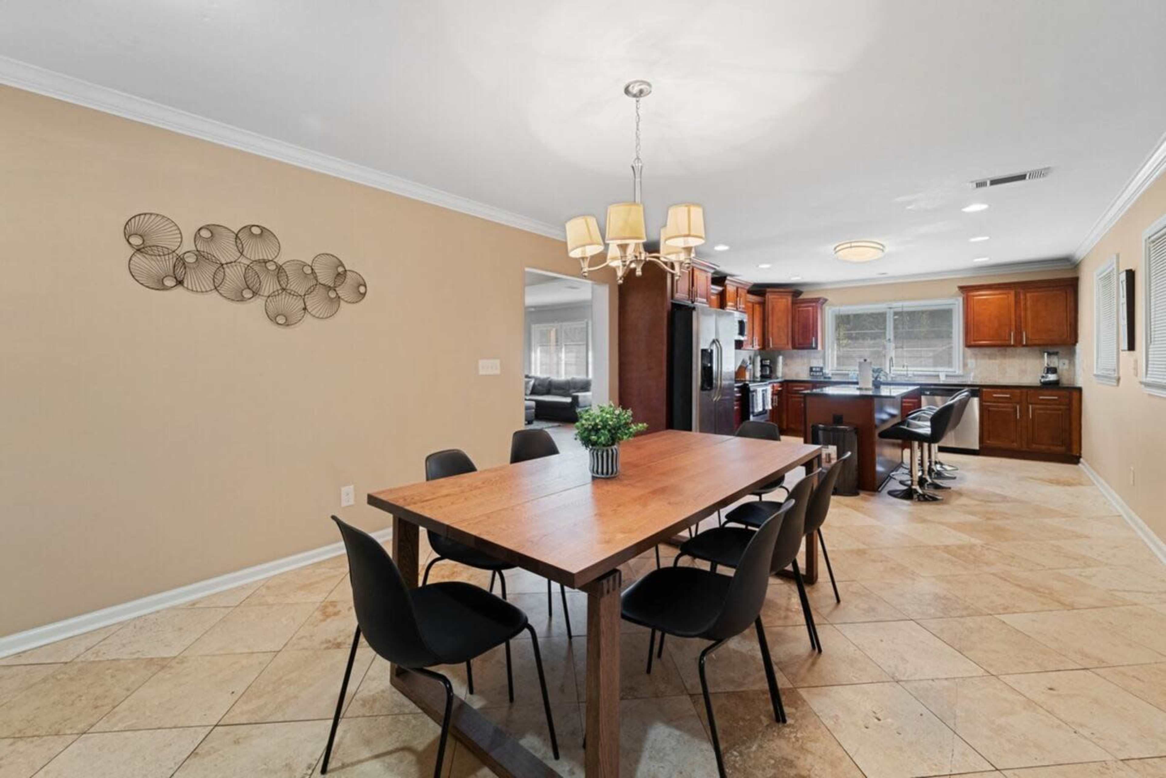 A dining area features a wooden table surrounded by black chairs, with a chandelier above and a view into a kitchen area in the background.