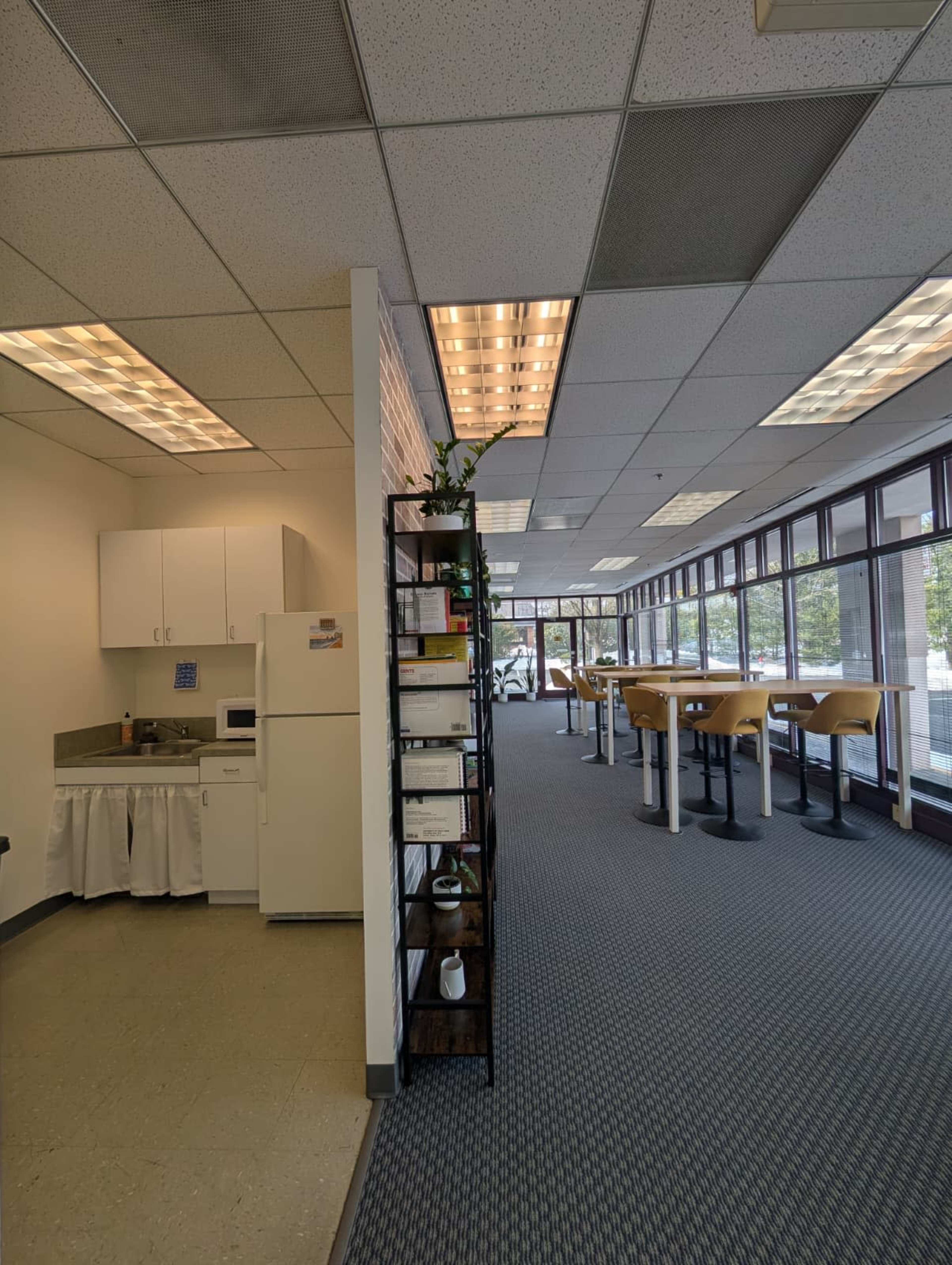 The image shows a spacious office area featuring a kitchenette on one side and tall tables with stools along a windowed wall.