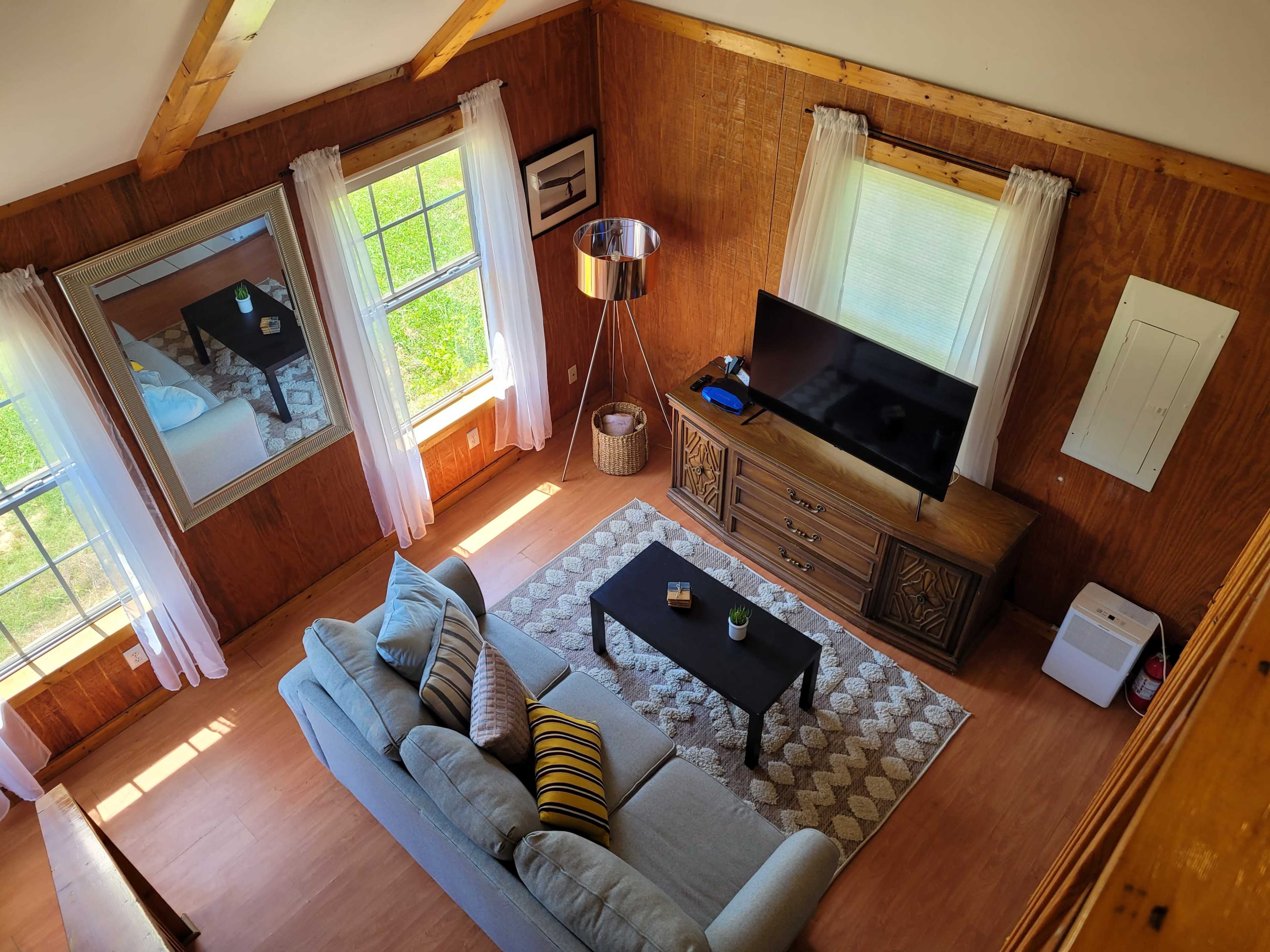 A cozy living room features a light blue sofa, a coffee table, and a television on a wooden cabinet, with large windows letting in natural light.