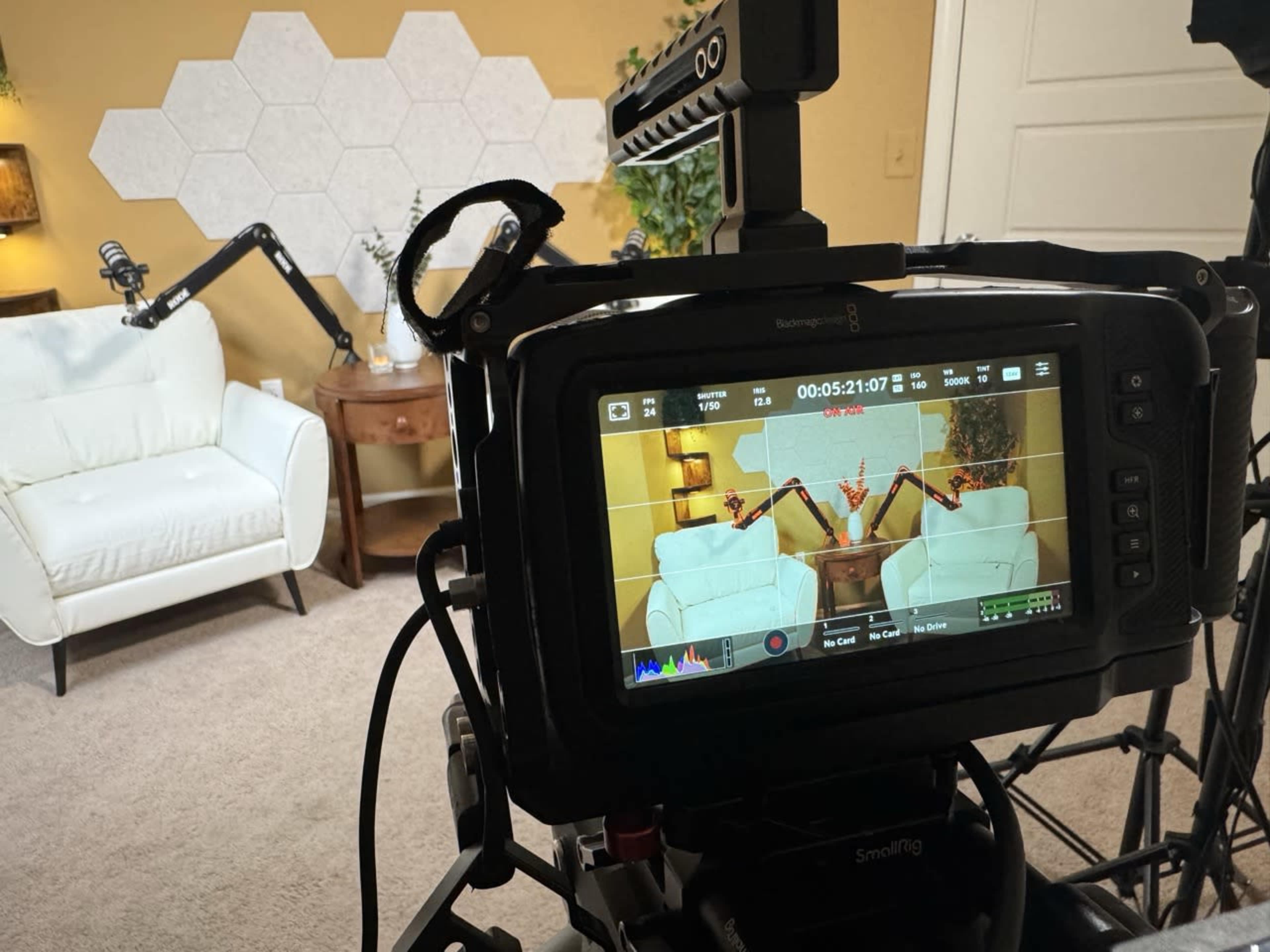 The image shows a video recording setup, featuring a white couch with decorative pillows, a side table, and a camera display showing the scene being filmed.