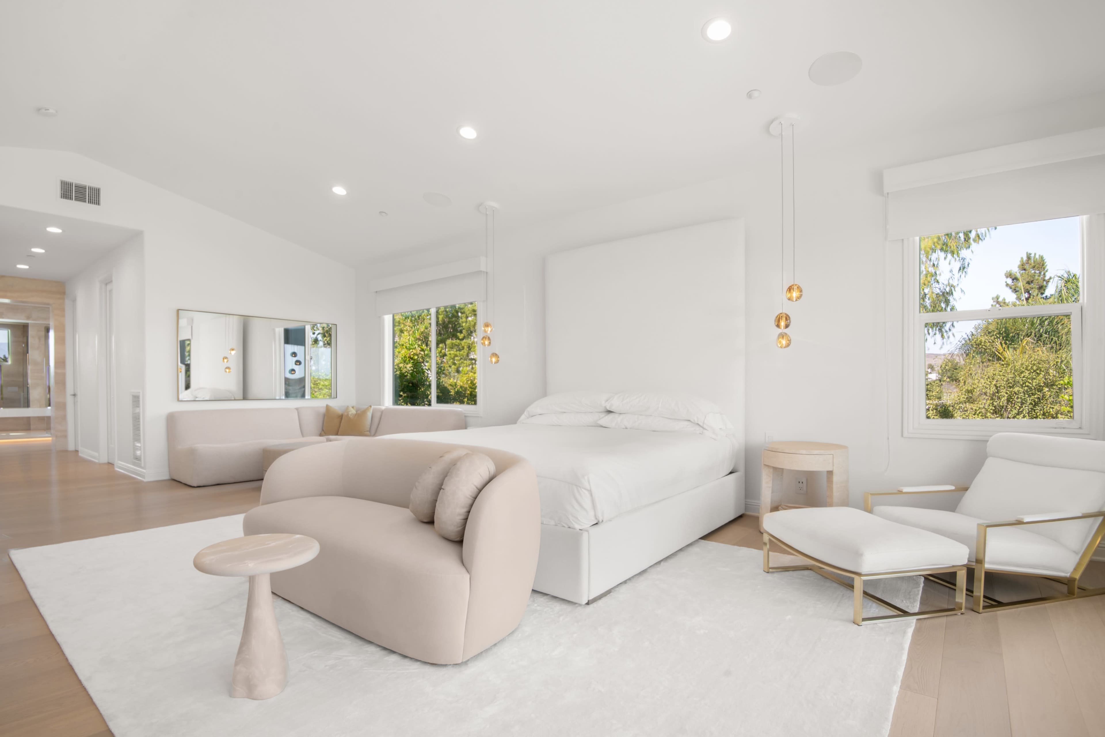 A minimalist bedroom features a bed with white bedding, a light-colored sofa, a small side table, and large windows offering a view of greenery.