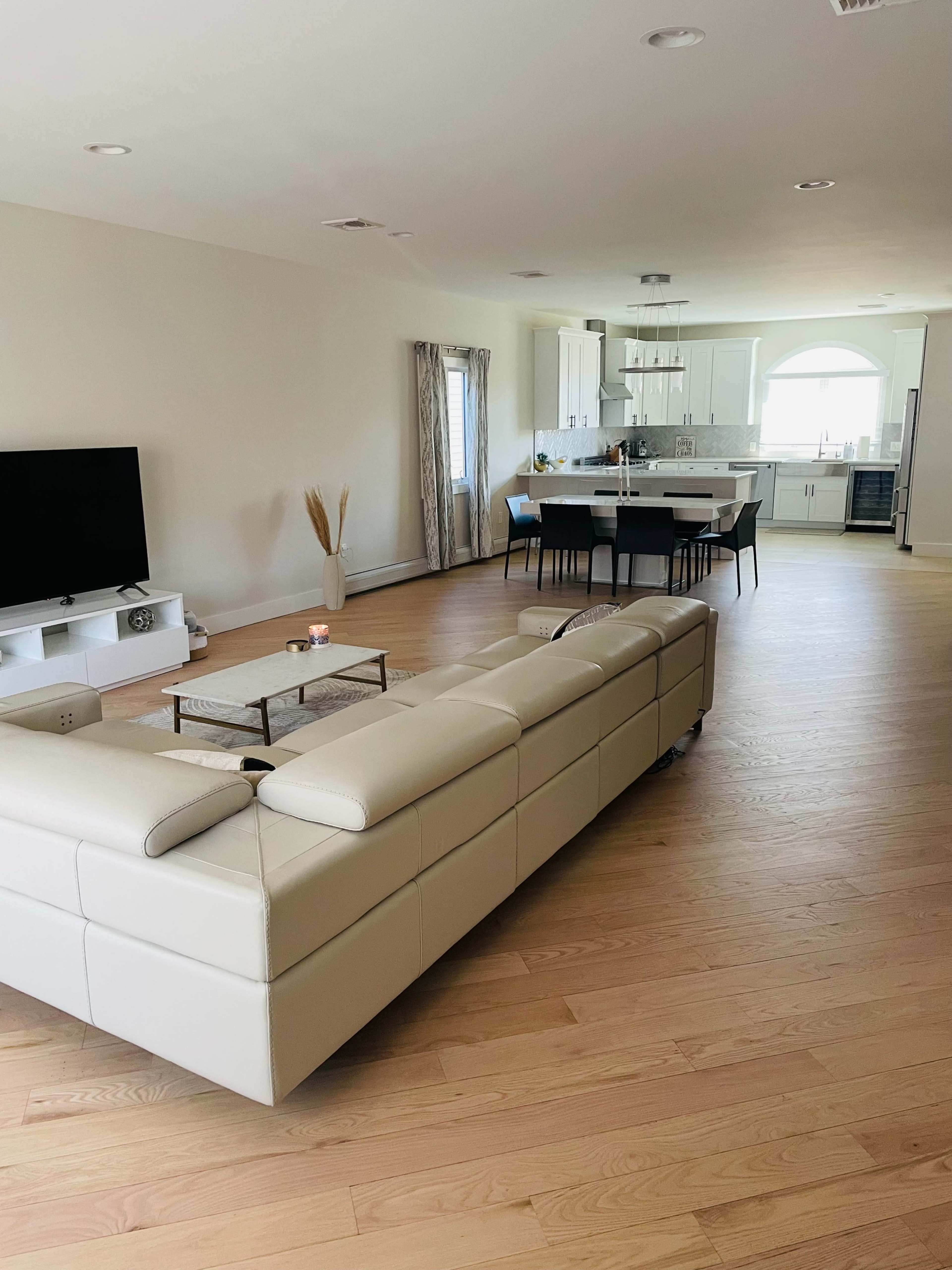 A spacious living area with a cream-colored sectional sofa facing a television, leading into an open kitchen with modern appliances and a dining table.