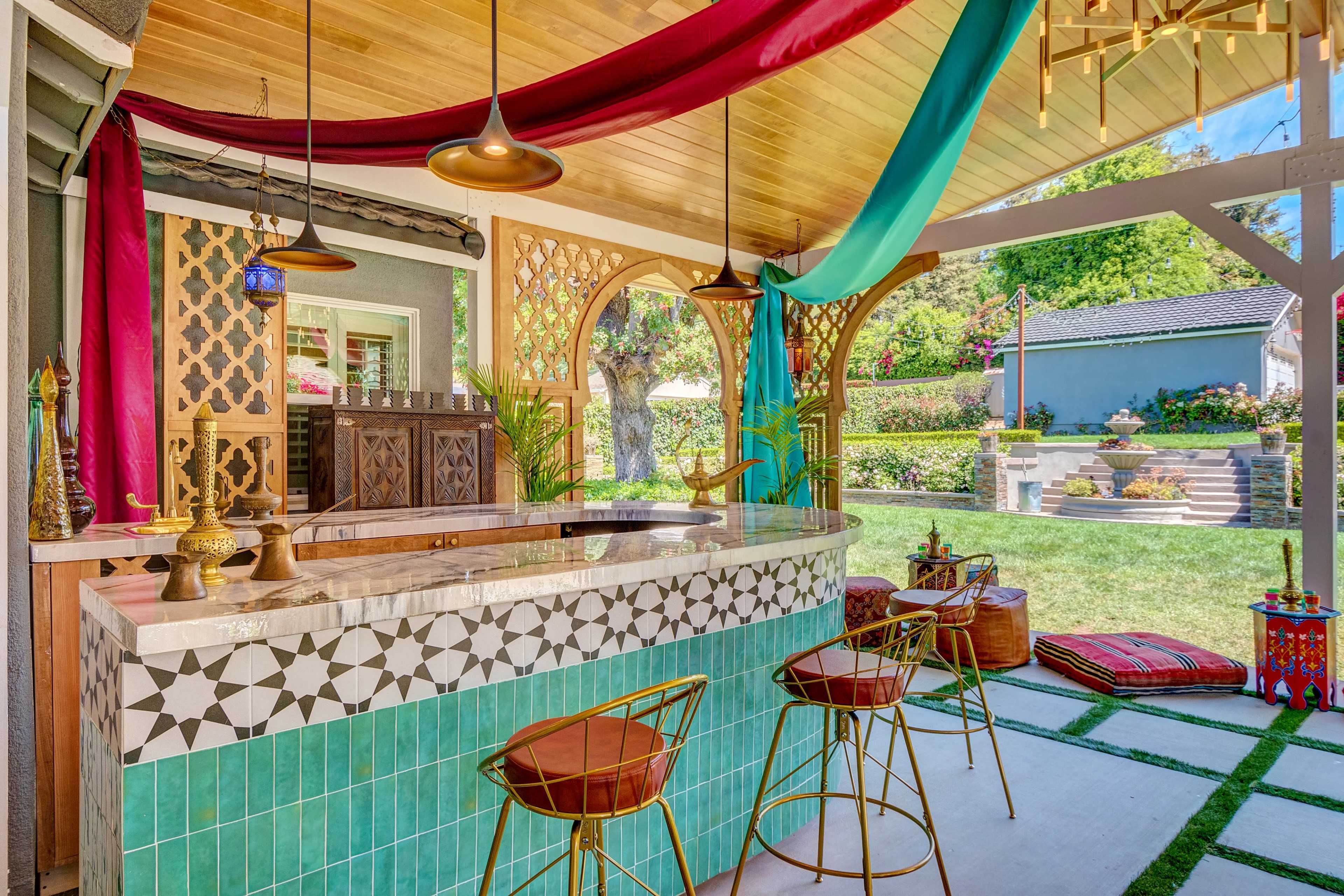 A vibrant outdoor bar area features a tiled counter with metallic stools, surrounded by colorful drapes and decorative elements, set against a lush garden backdrop.