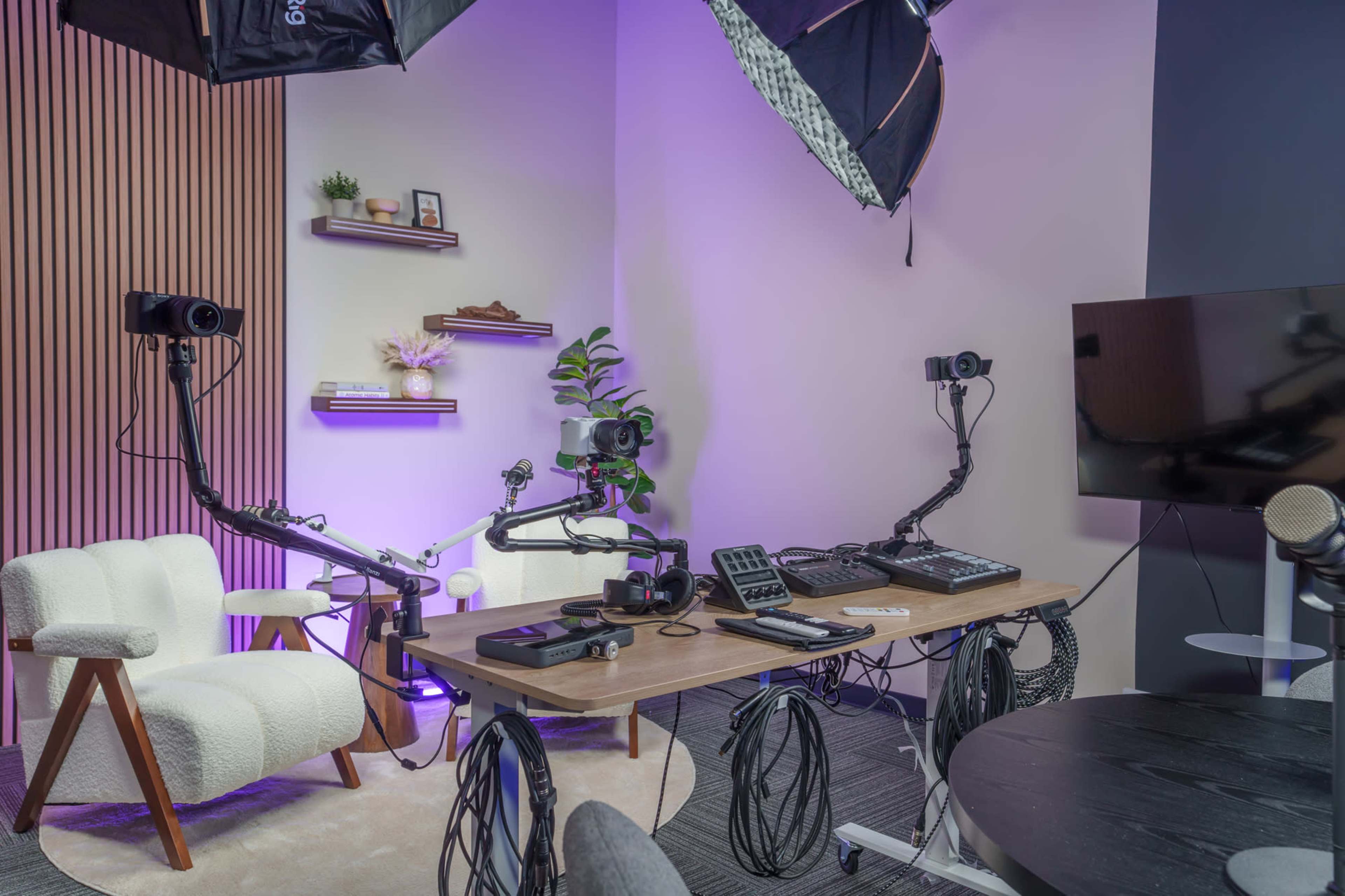 The image shows a modern studio setup featuring two cameras on articulated mounts, a wooden table with various equipment, and a comfortable seating area against a backdrop of wood paneling and decorative shelves.