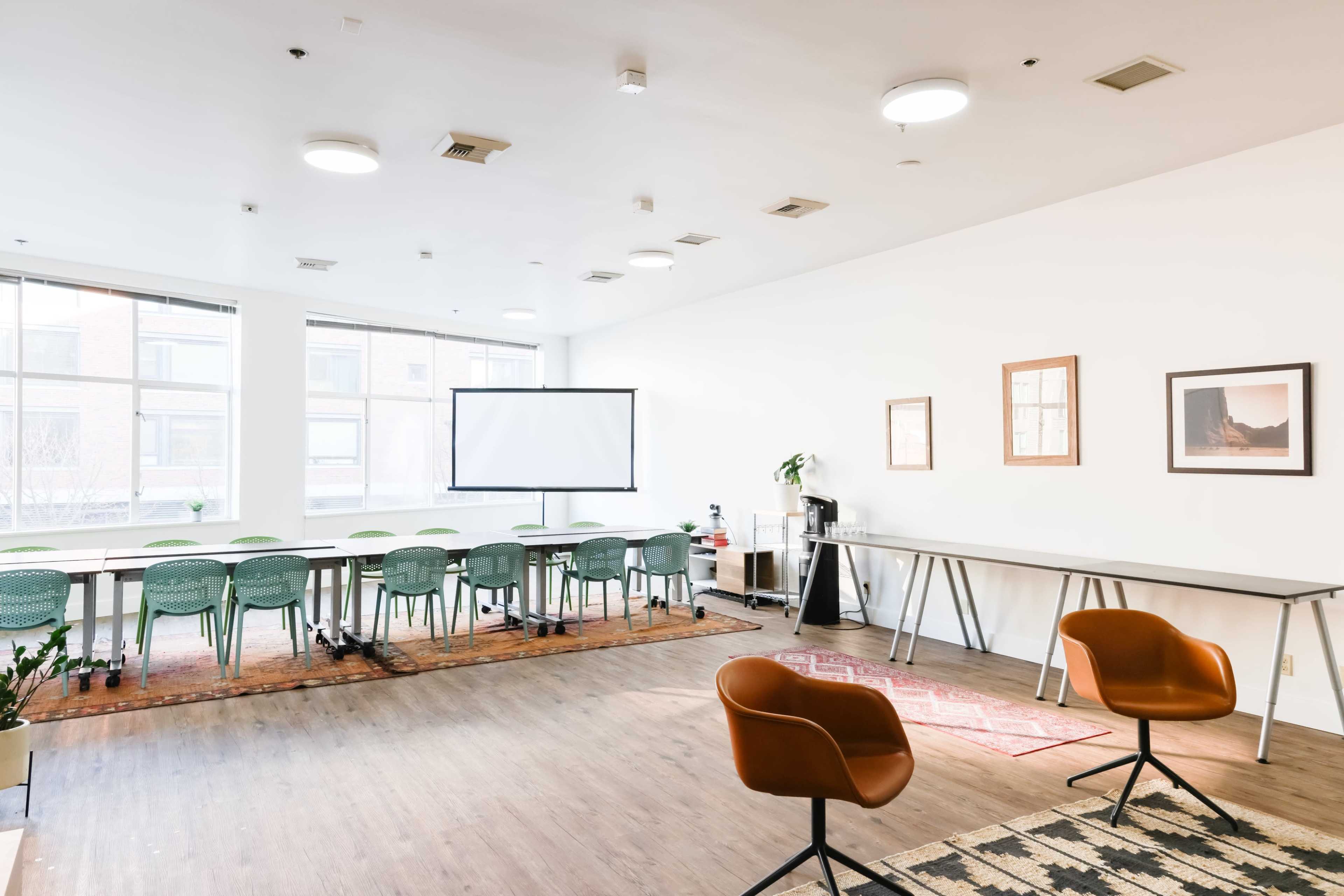 A brightly lit conference room features a long table with green chairs, a projector screen, and two brown chairs positioned in front.