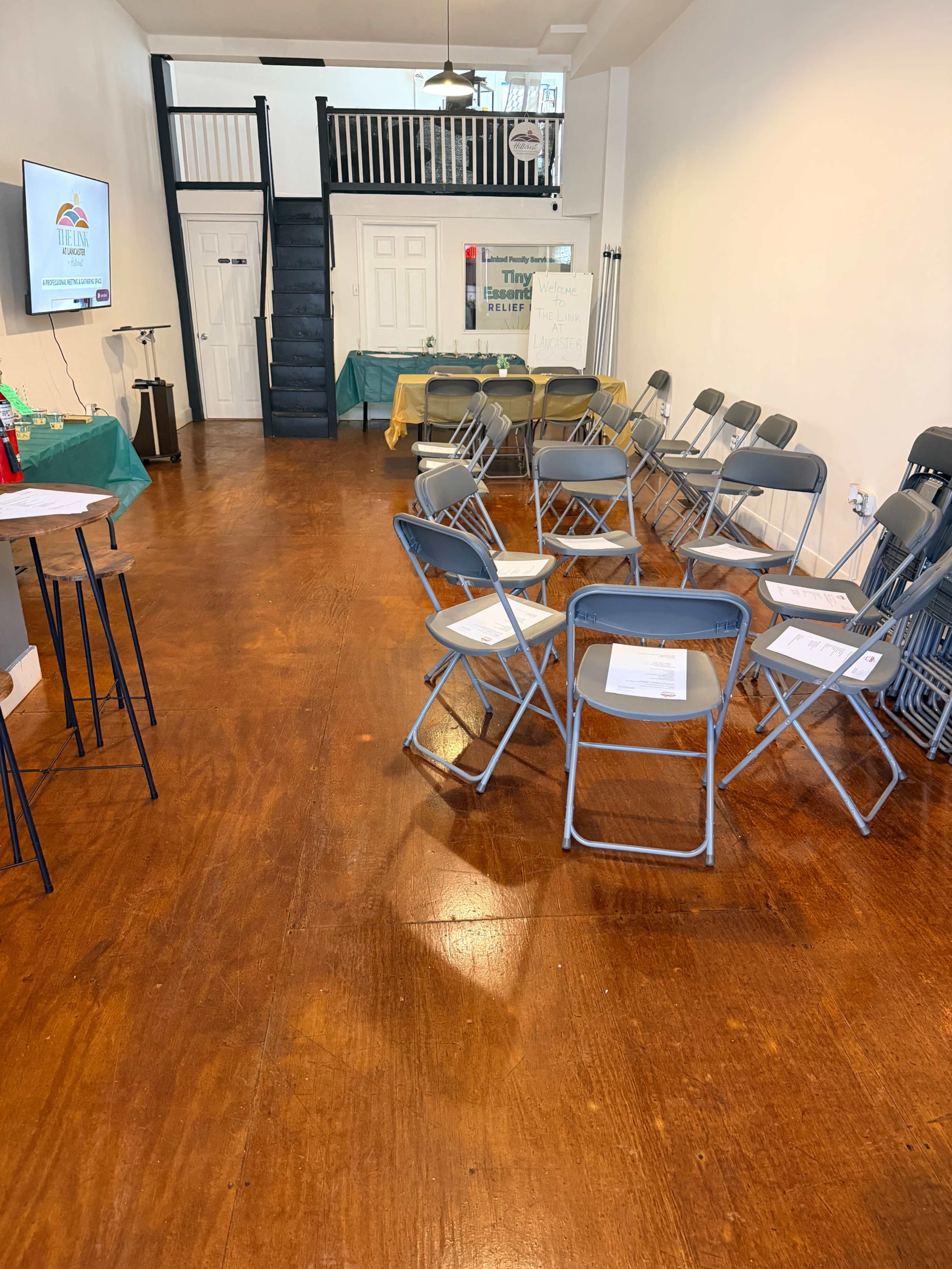 The image shows a room with a polished wooden floor, featuring a set of folding chairs arranged in a semi-circle and a staircase leading to an upper level.