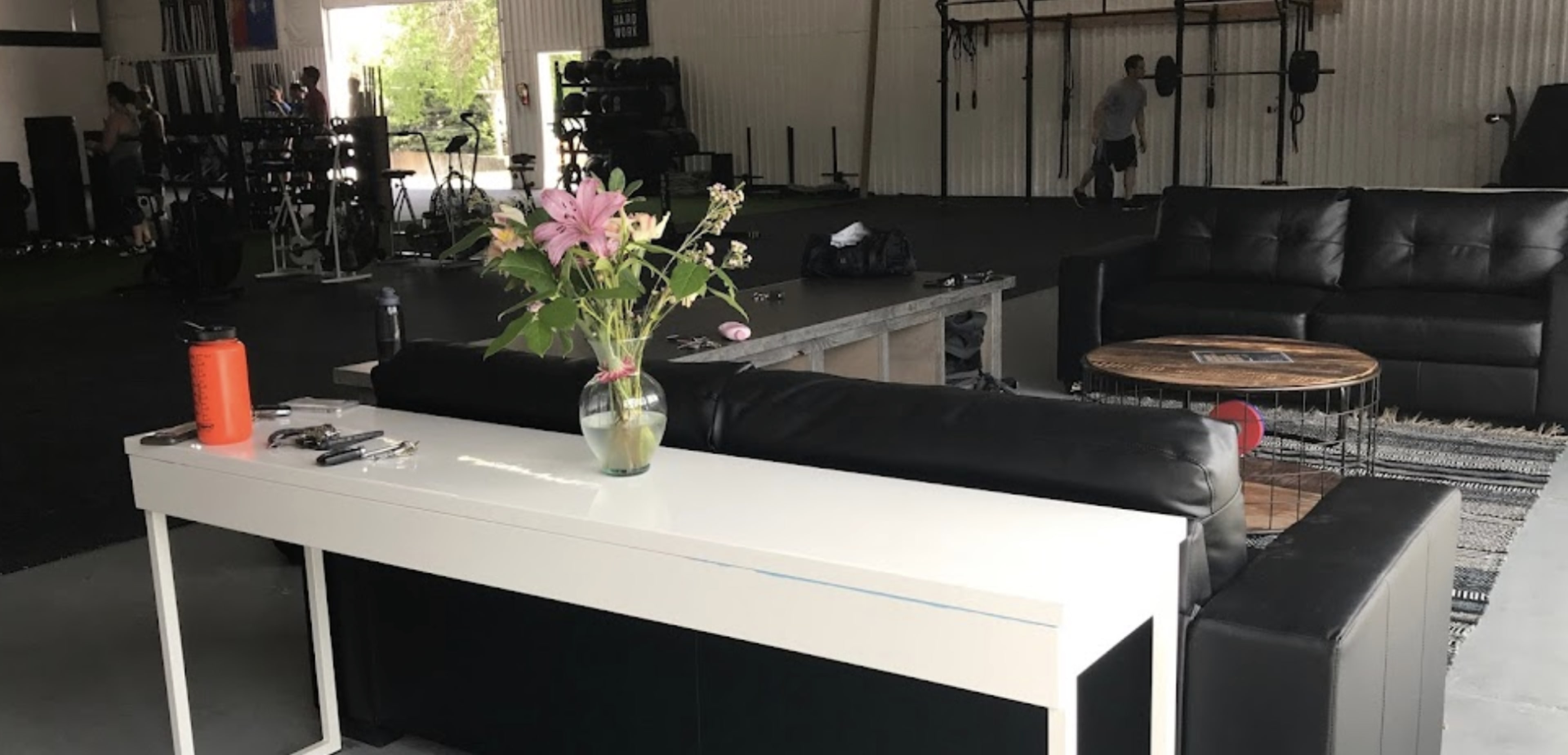The image shows a spacious gym interior with black workout equipment, a seating area featuring a black couch, coffee table, and a vase of flowers on a white console table.