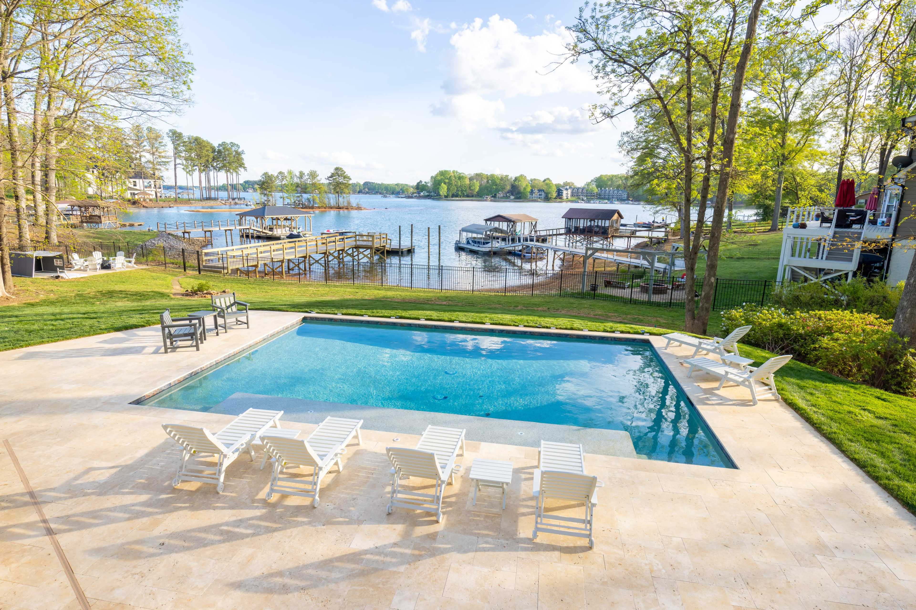 A clear swimming pool with white lounge chairs surrounds it, overlooking a lake with several docks and boats under a bright sky.