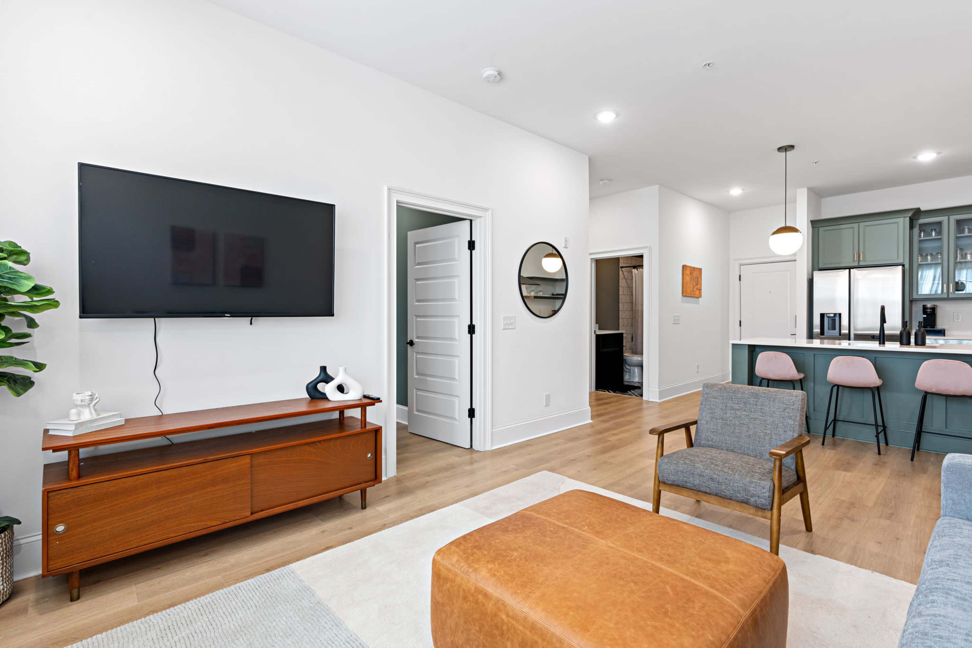 A modern living room features a wall-mounted television, a wooden entertainment center, a gray armchair, an ottoman, and an open kitchen area in the background.