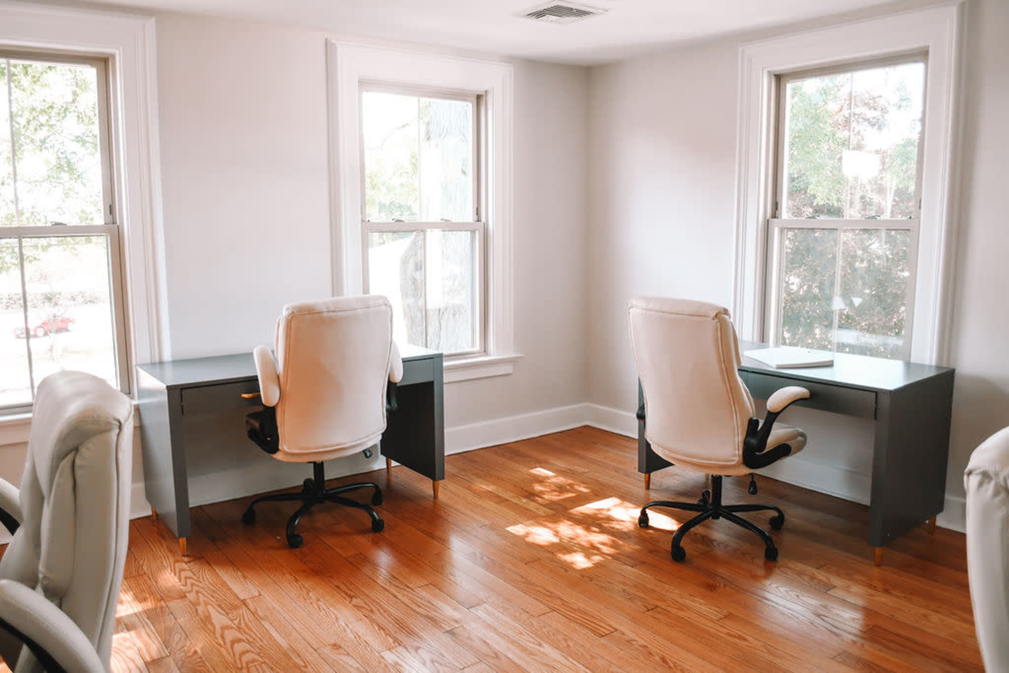 The image shows a bright room with two desks and chairs positioned near windows, featuring hardwood flooring.