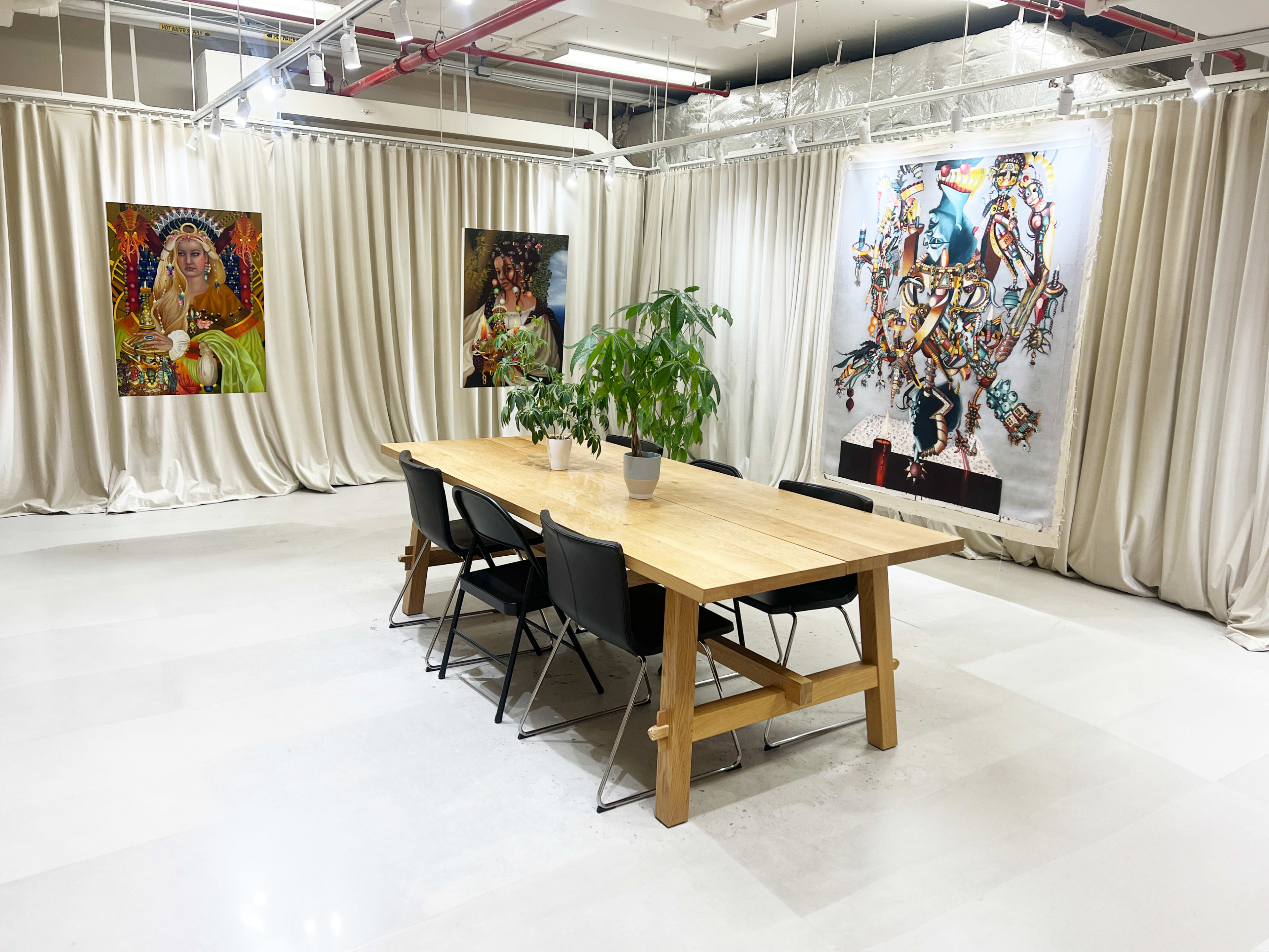 A spacious meeting room features a large wooden table surrounded by black chairs, with colorful artwork displayed on the walls behind floor-to-ceiling curtains.