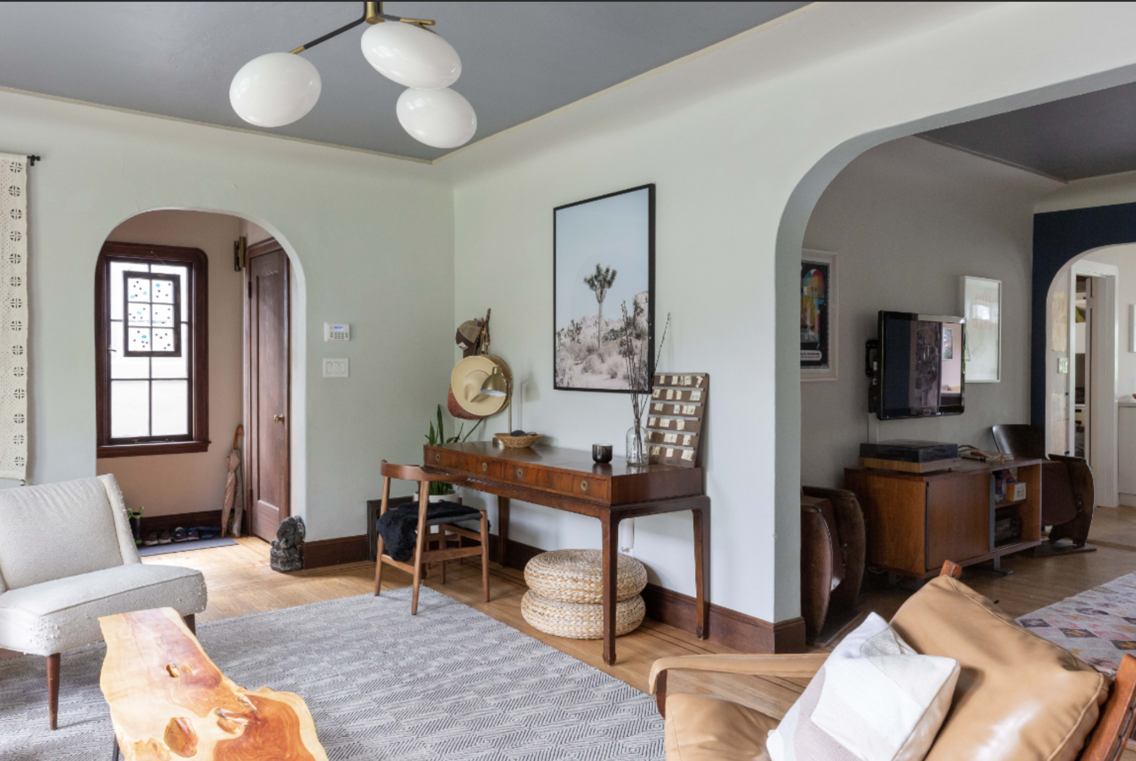 A modern living room featuring a wooden table, a light fixture with three circular lamps, and various decorative elements against a softly colored wall.