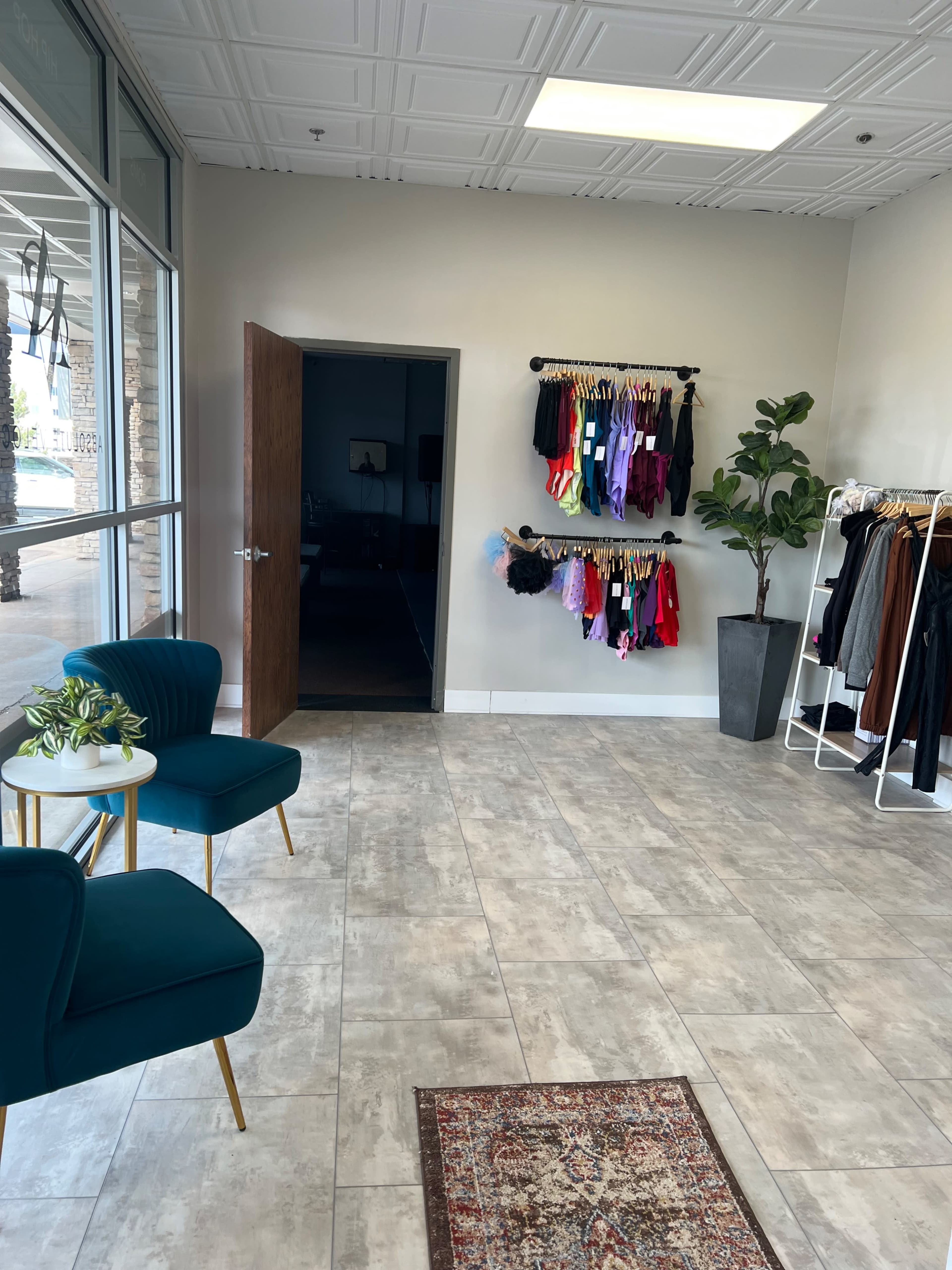 A retail store interior with two teal chairs and a small table on one side, a clothing rack with colorful garments, and an open door leading to another room.