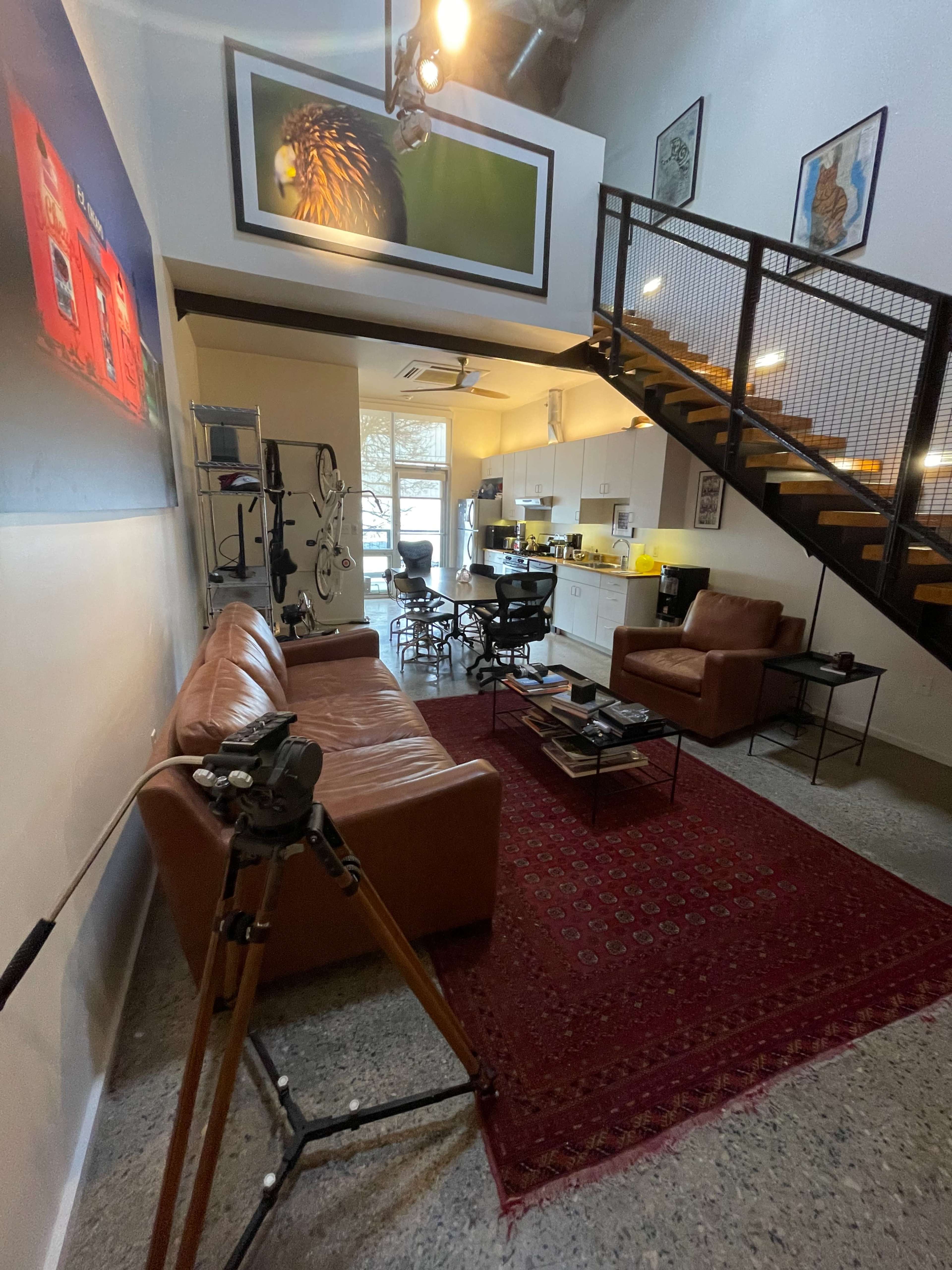 A spacious living area with a brown leather sofa, a dining table, and a staircase leading to an upper level, featuring large windows that allow natural light to enter.