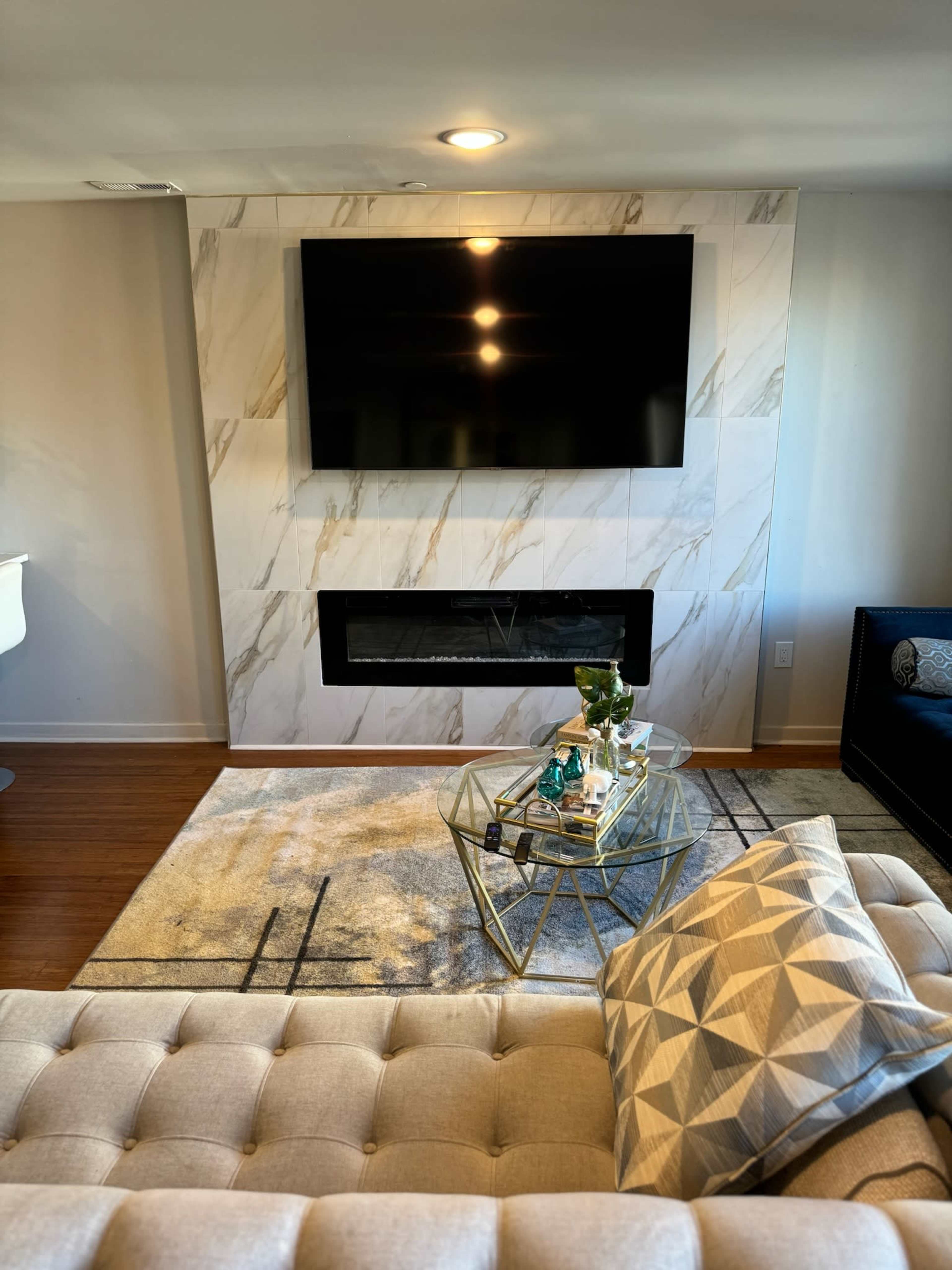 The image shows a modern living room with a wall-mounted TV above a fireplace, a glass coffee table with decorative items, and a sofa with a patterned cushion.