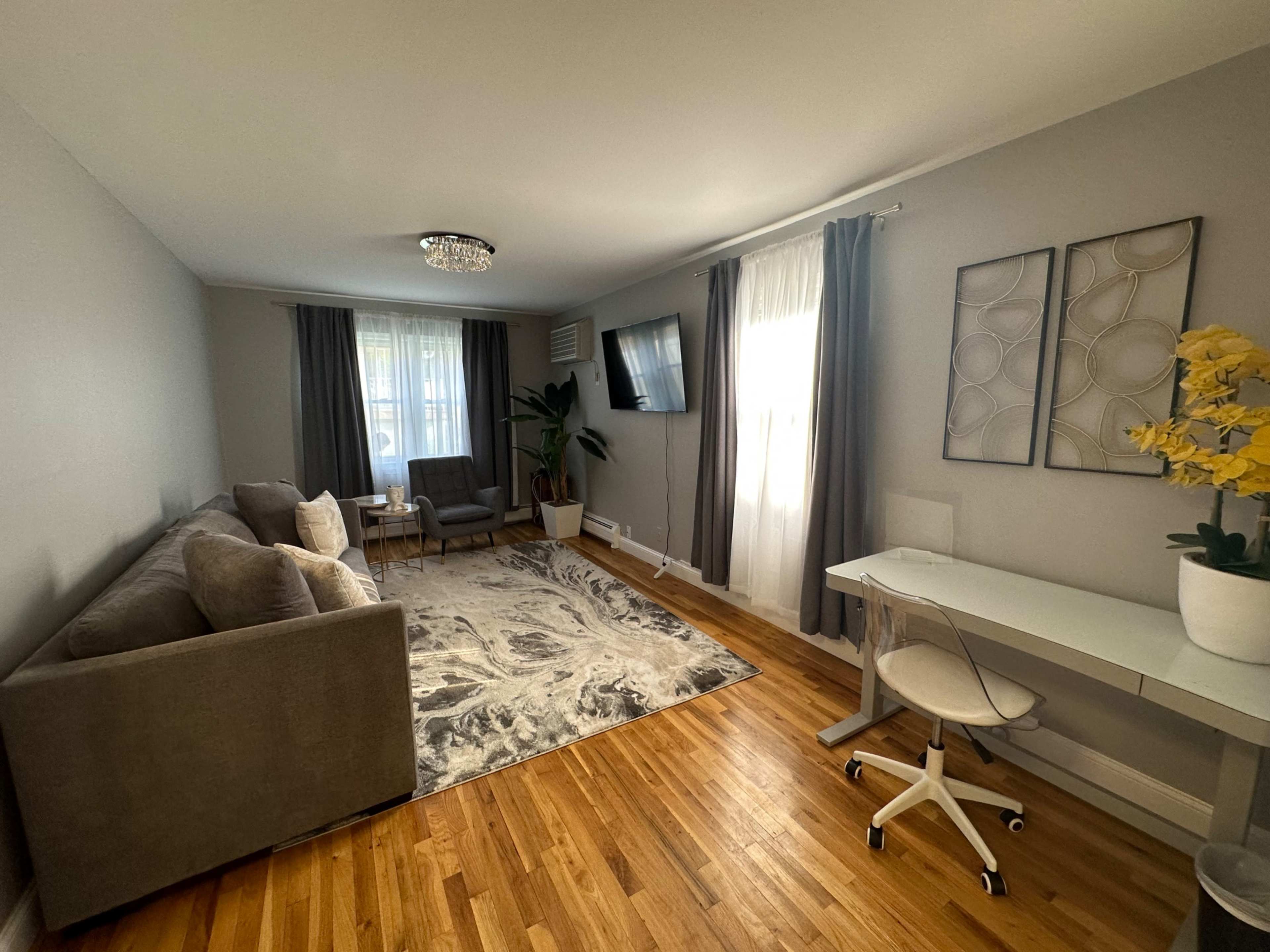 A contemporary living room with a gray sofa, two chairs, a coffee table, a wall-mounted TV, and a small office desk, all illuminated by natural light from large windows.