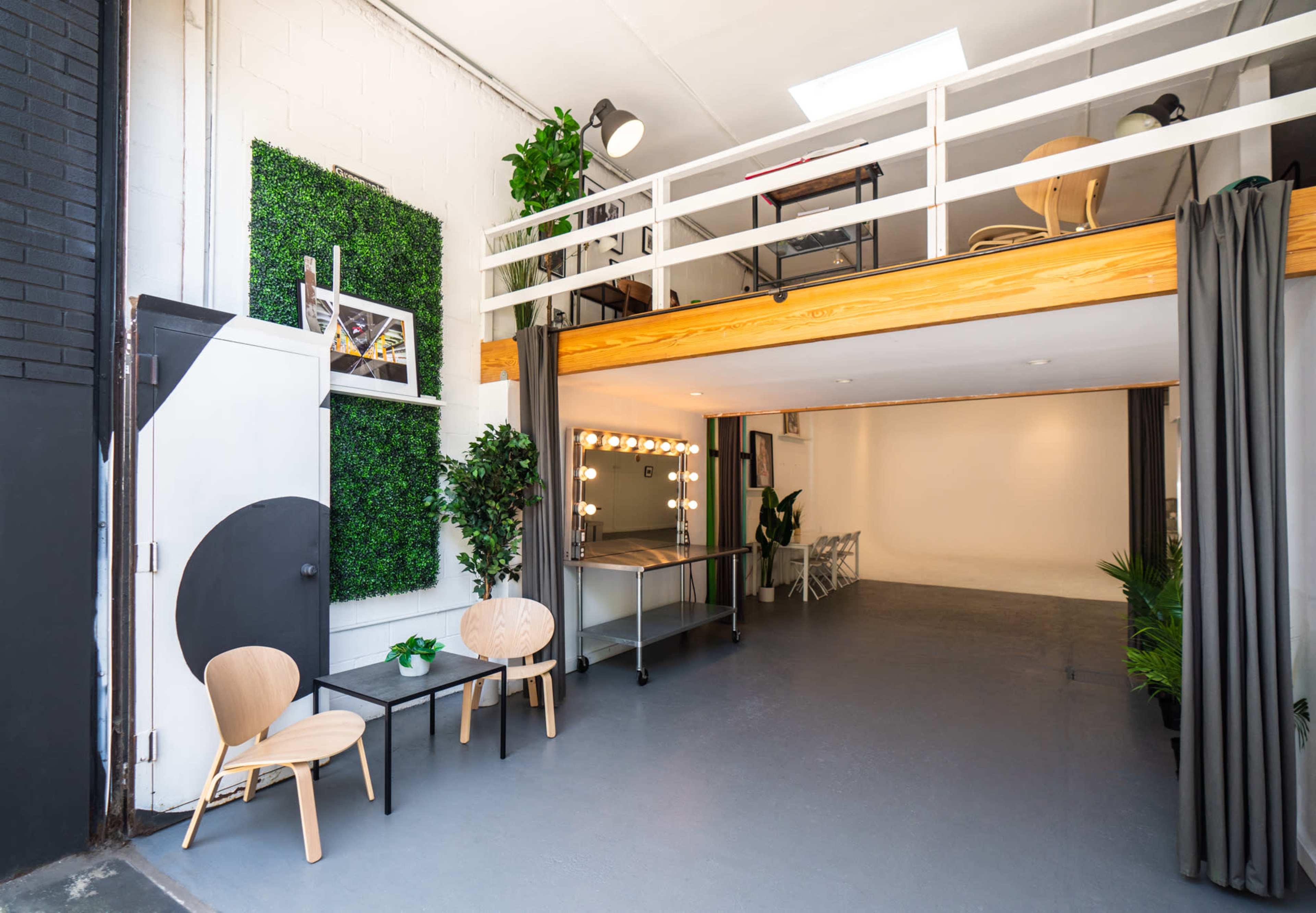 The image shows an interior space featuring a lofted area, a green wall installation, a seating arrangement with two chairs, and a lighted vanity mirror.