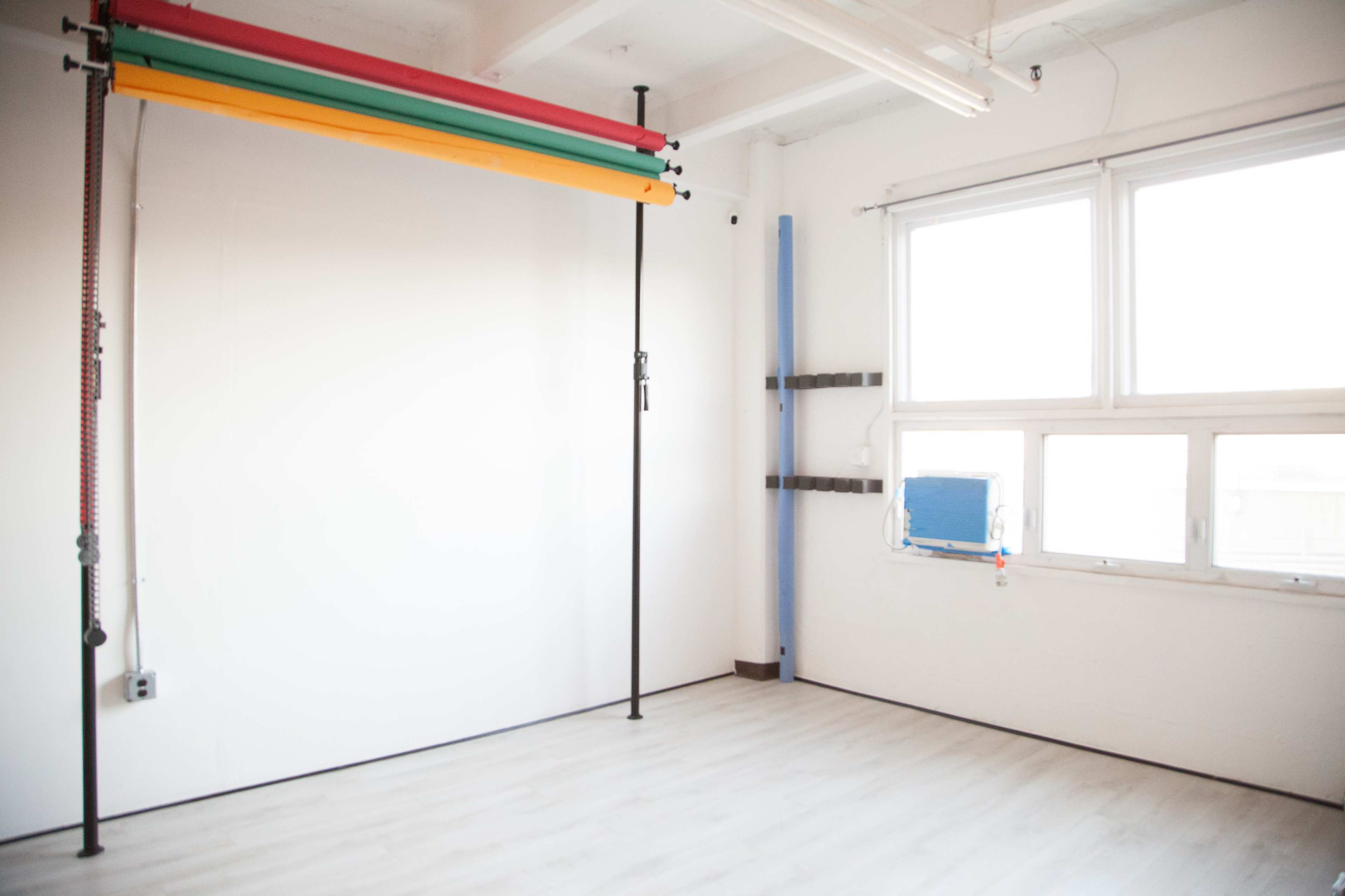 The image shows a bright, empty studio space with a blank white wall and colorful backdrops hanging on a vertical support.