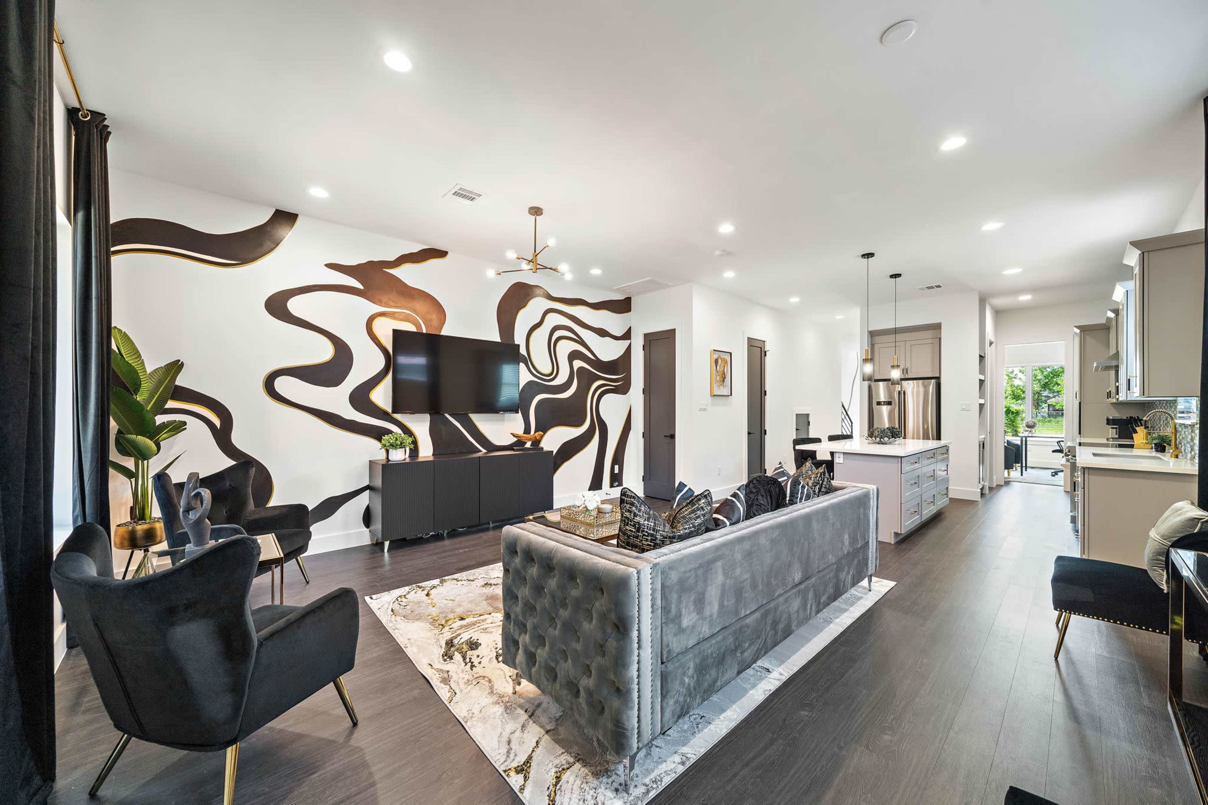 The image shows a modern living room with a sectional sofa, a large wall-mounted TV, and a decorative mural on one wall, leading into a kitchen area.