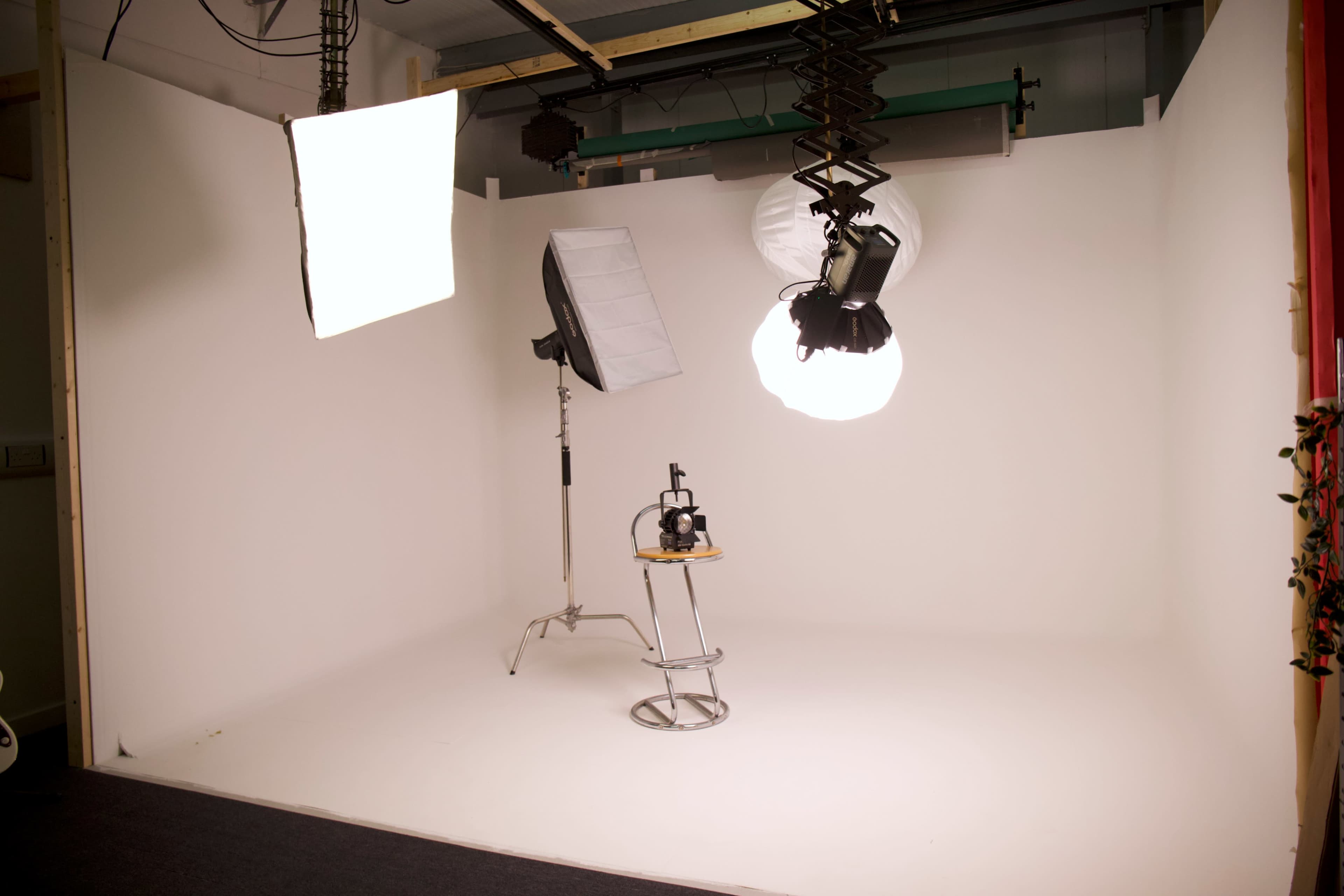 The image shows a photography studio with a white backdrop, multiple light sources, and a stool positioned for shooting.