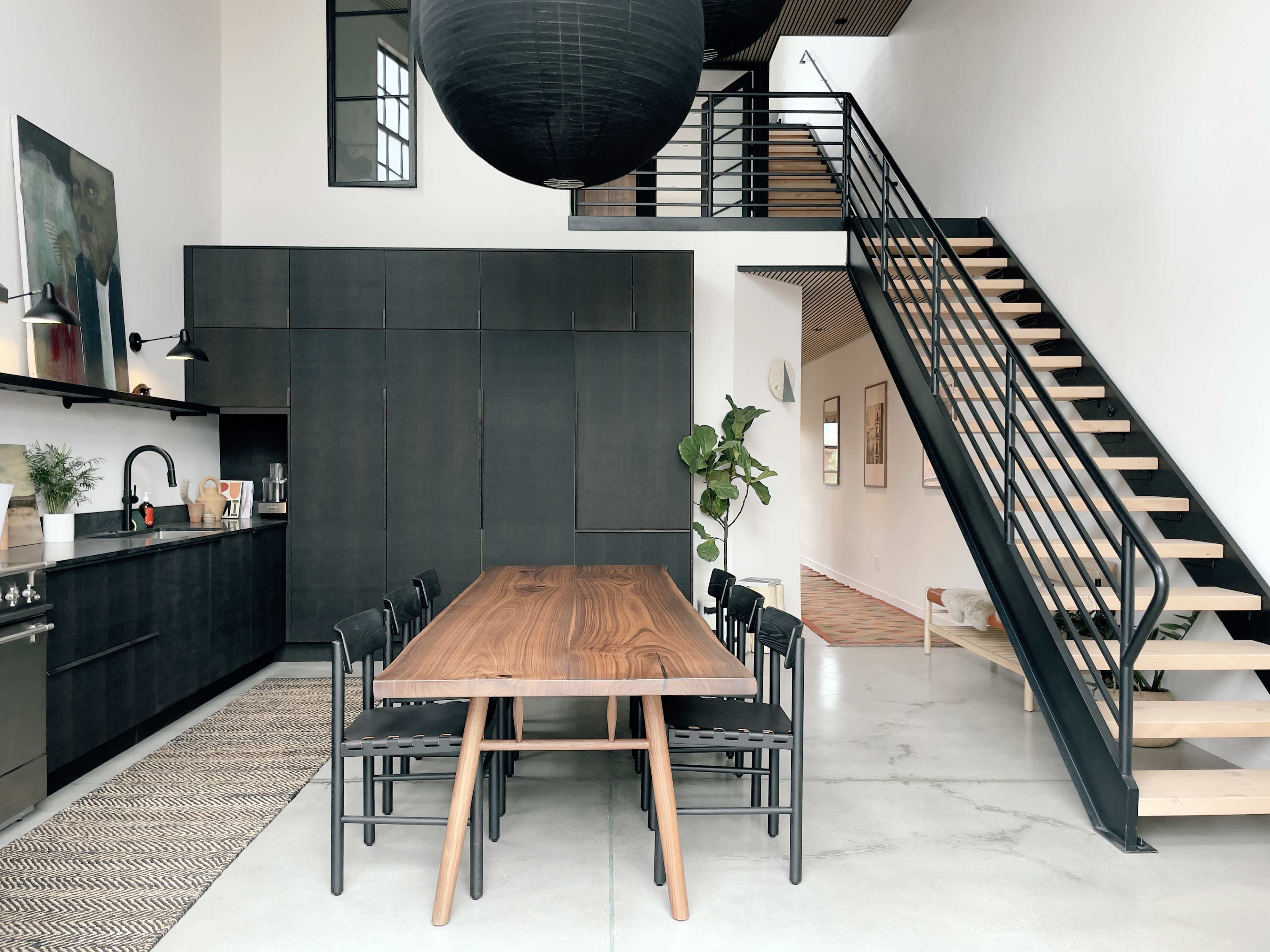 A modern kitchen and dining area with a wooden table surrounded by black chairs, set against a backdrop of dark cabinetry and a staircase leading to an upper level.