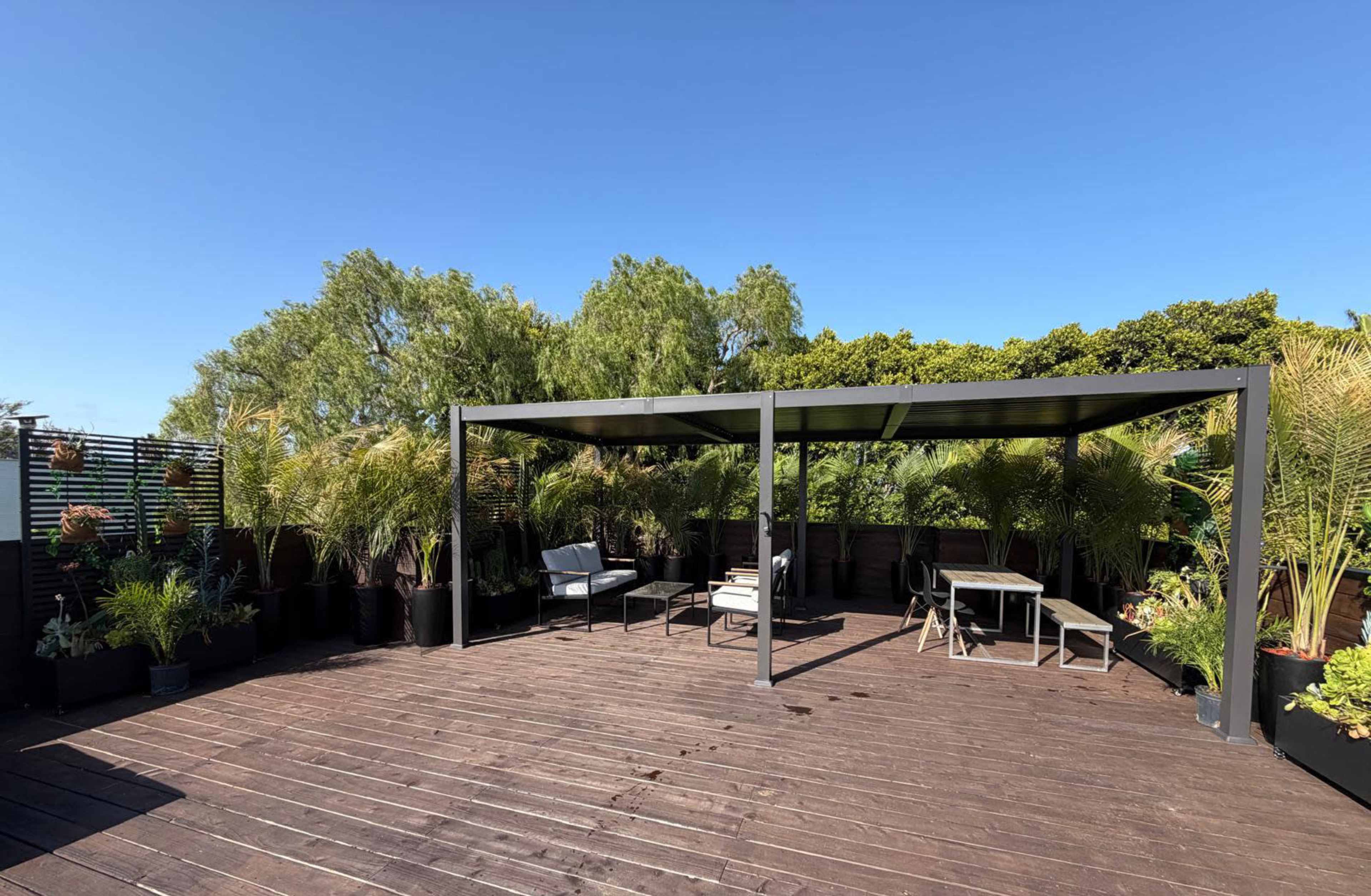 A rooftop terrace features a shaded seating area with a table and chairs, surrounded by potted plants and trees under a clear blue sky.