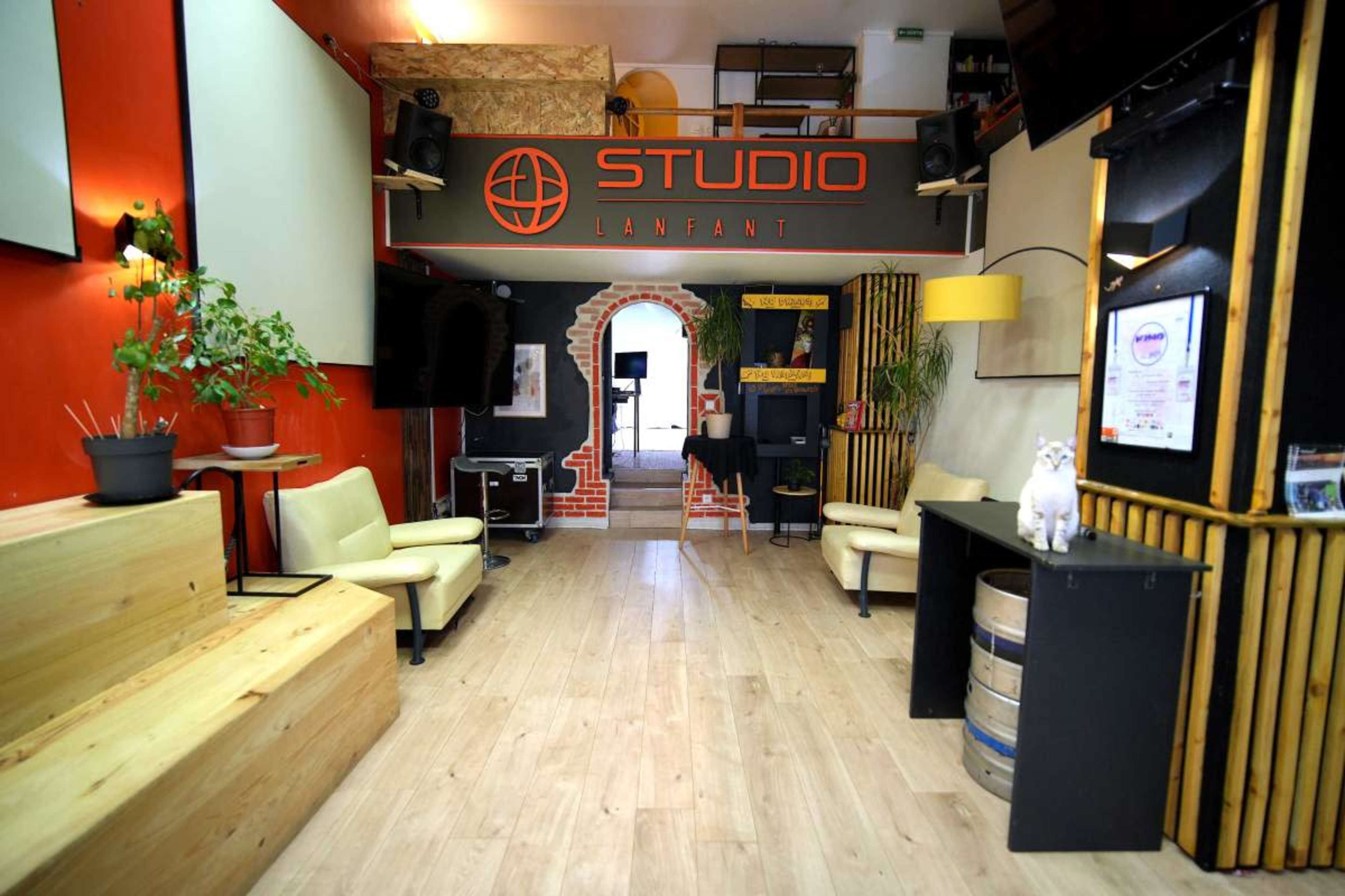 The image shows a studio interior with wooden flooring, a red accent wall, and a sign labeled "STUDIO LANFANT," featuring couches and various furnishings.