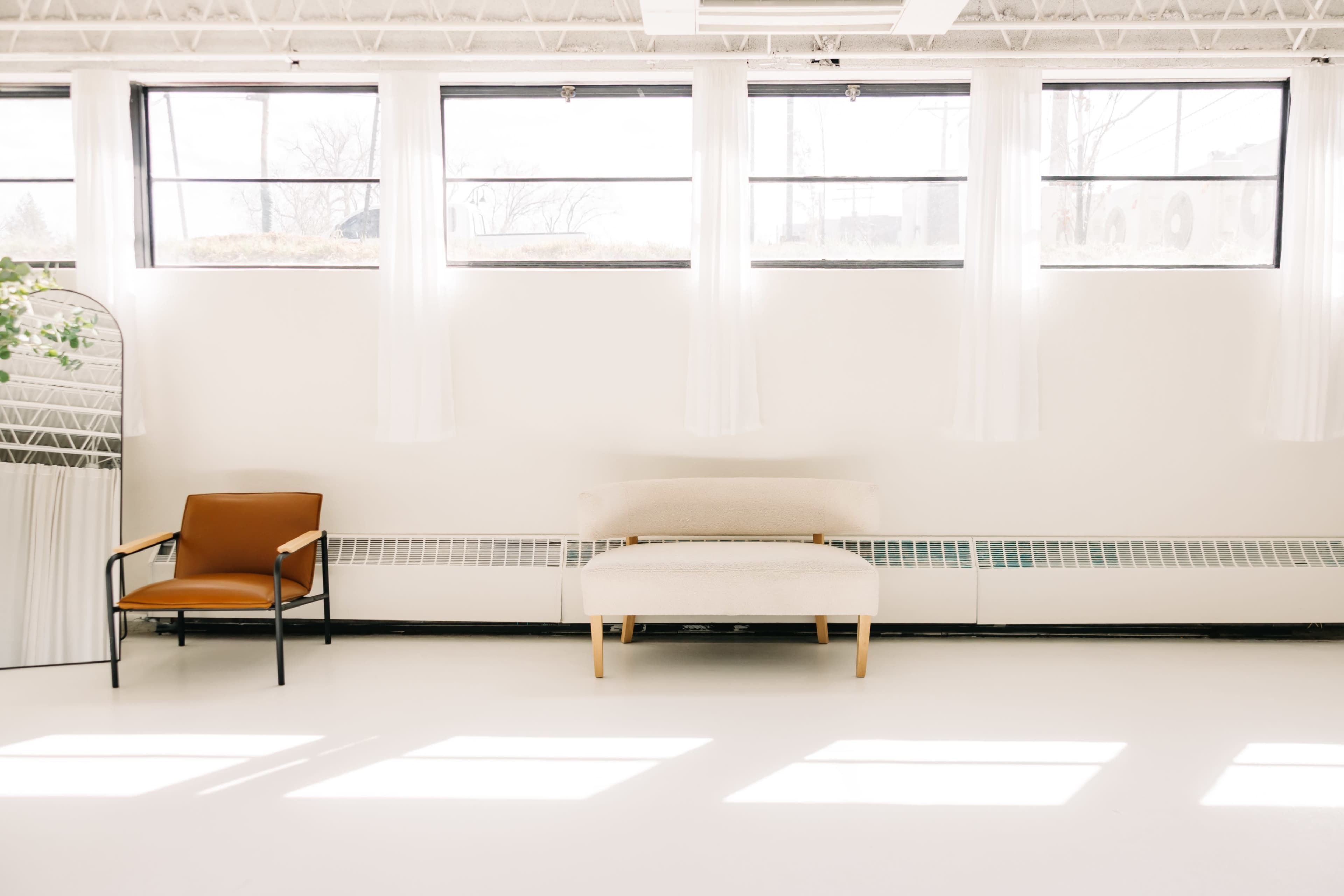 The image shows a bright, minimalist room with large windows, a white floor, a beige sofa, and a brown armchair.