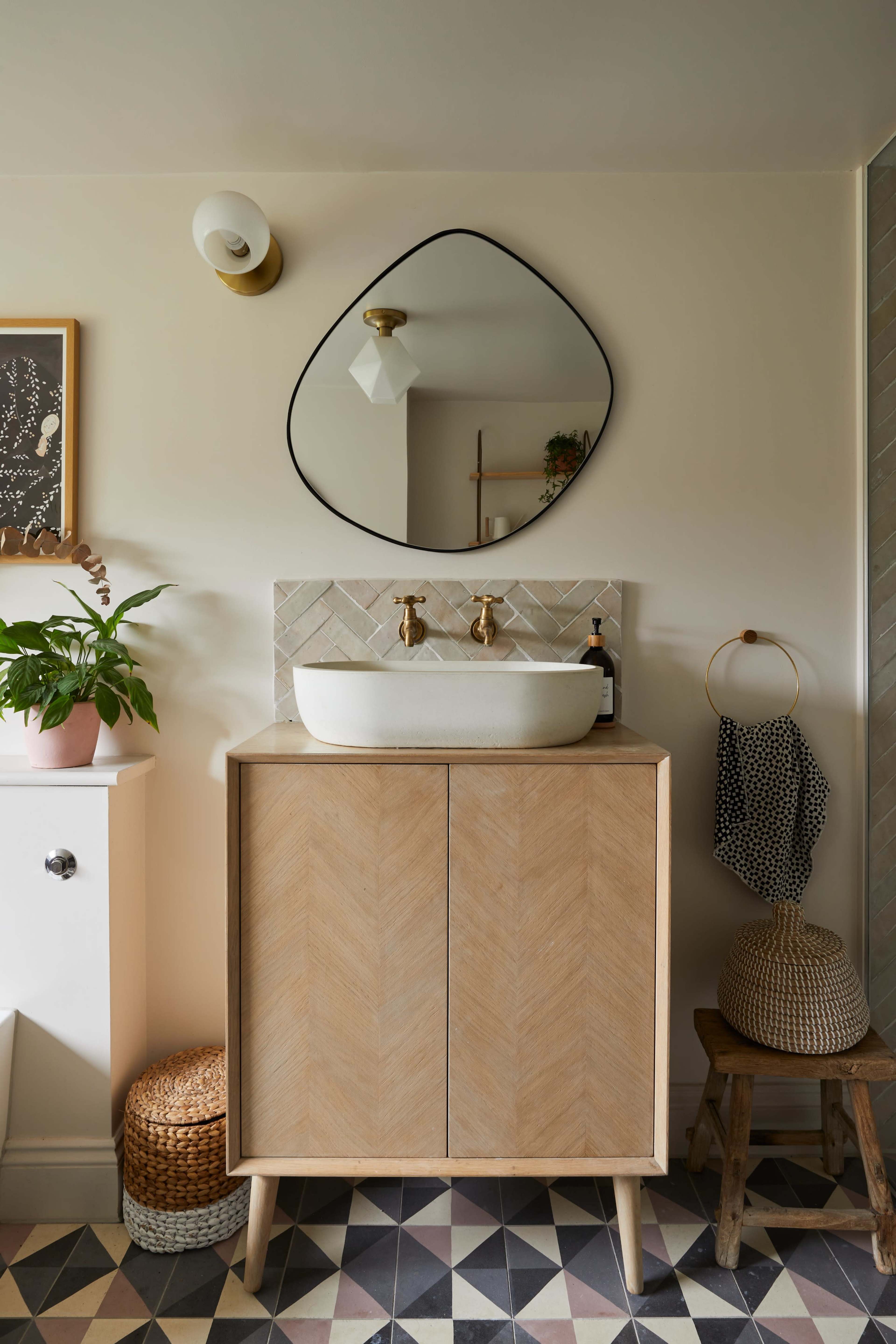 A bathroom features a wooden cabinet with a white basin on top, alongside a decorative mirror, plants, and patterned floor tiles.