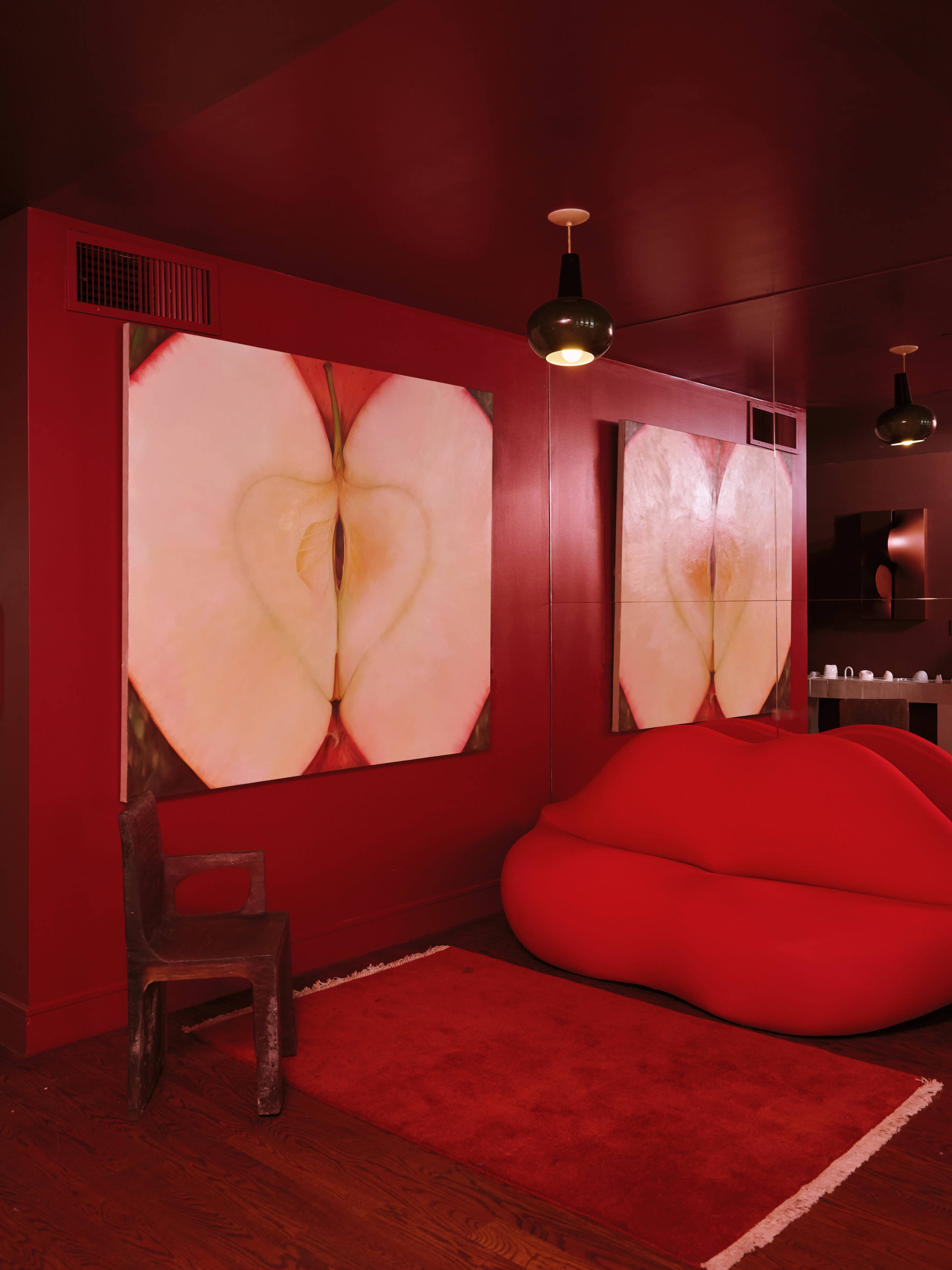 The image shows a modern room with red walls, featuring two large apple-themed artworks and a lip-shaped red sofa.