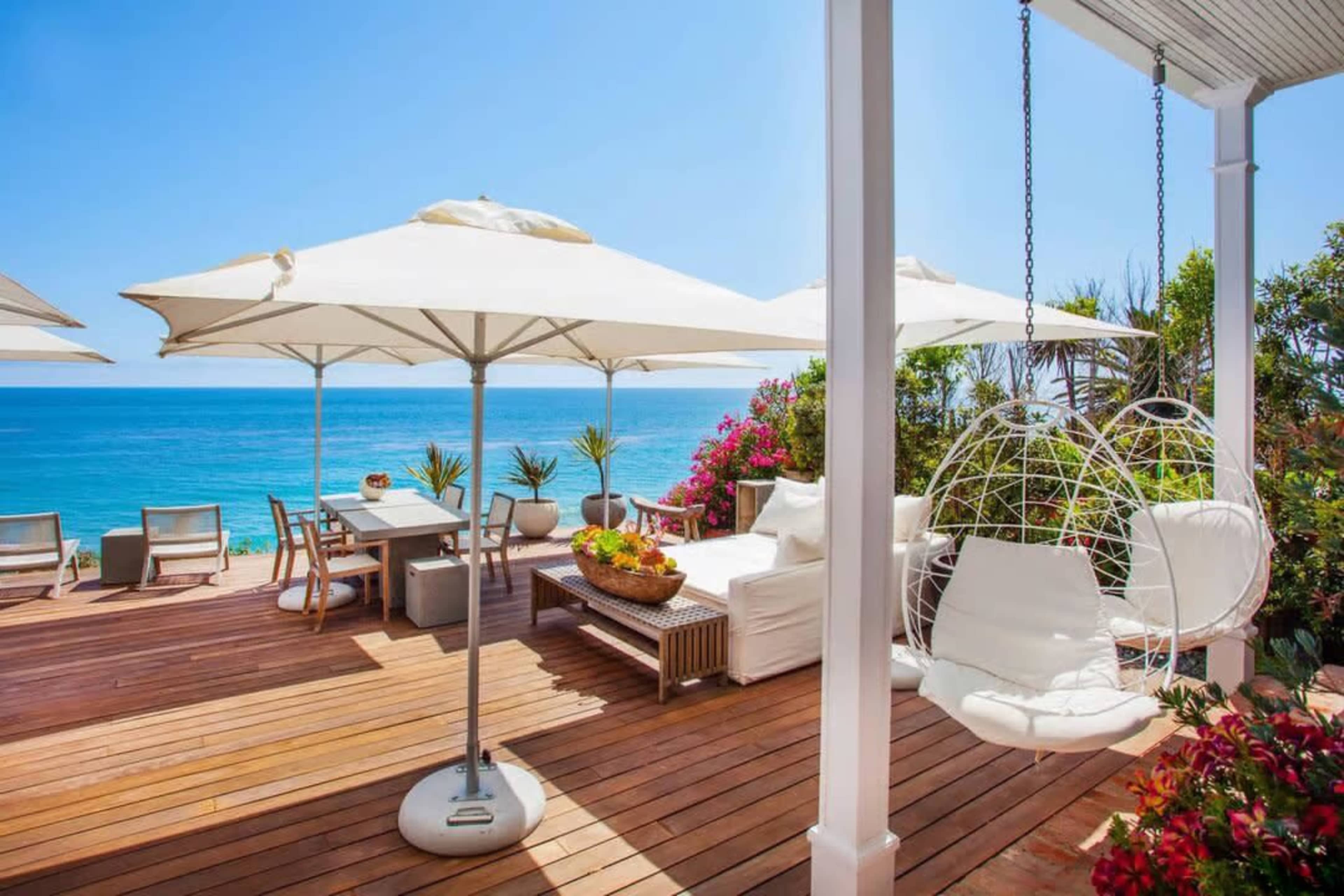 A seaside deck features a dining area with a table and chairs, large umbrellas, potted plants, and a hanging chair overlooking the ocean.
