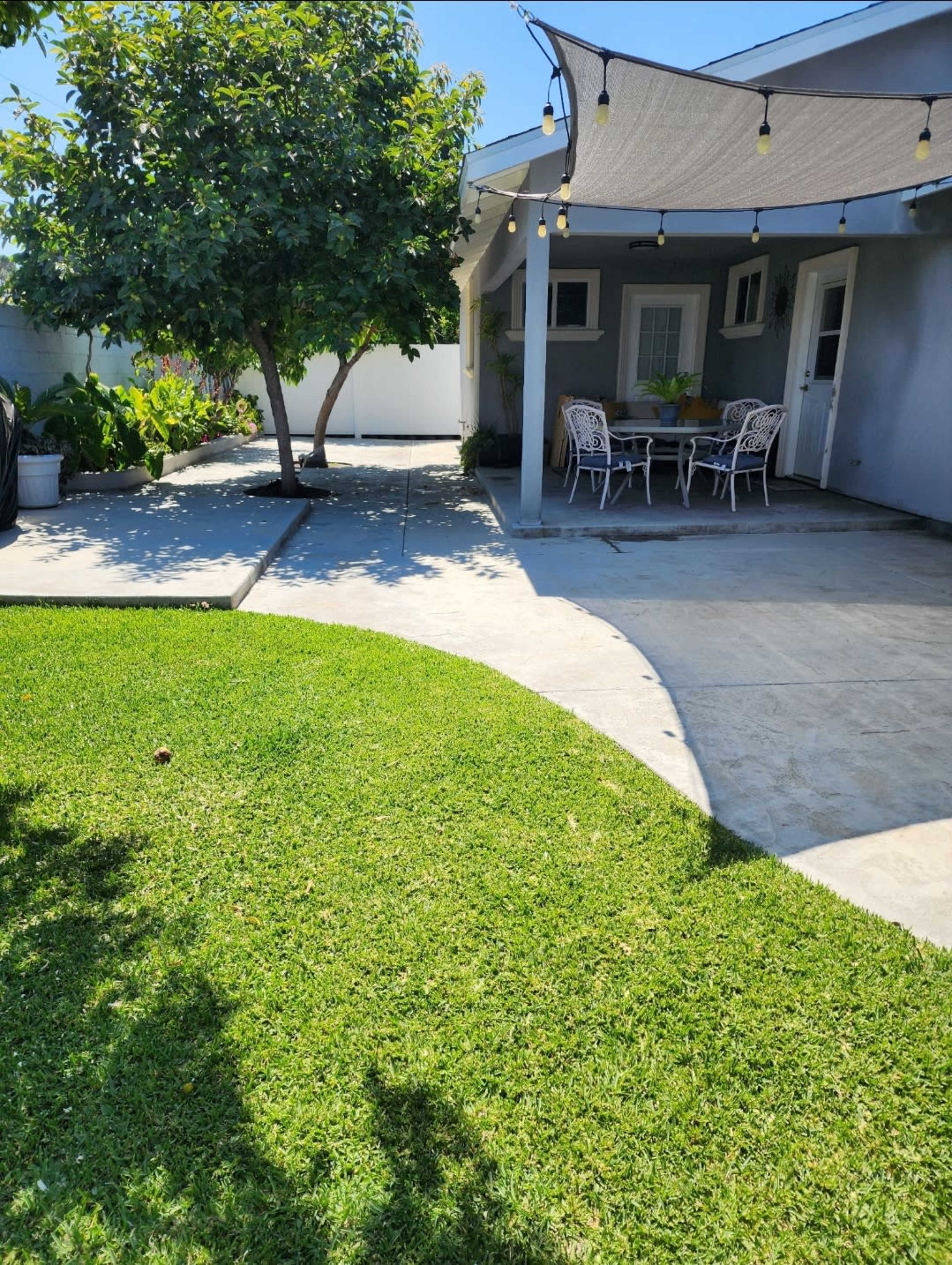 The image shows a backyard with a neatly cut lawn, a patio area with a table and chairs, and a tree providing shade.