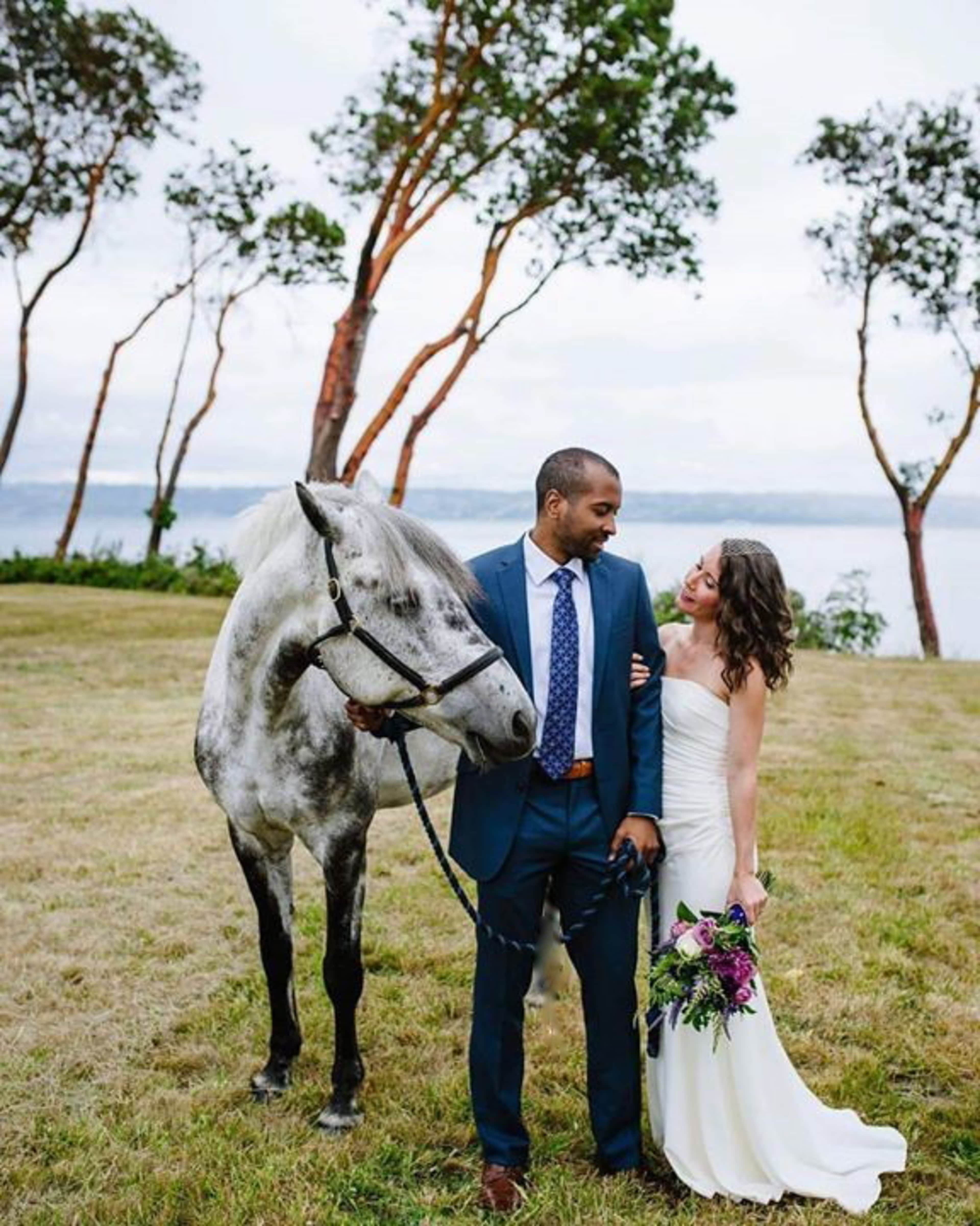 A couple stands together in a grassy area near a body of water, with a gray horse beside them.