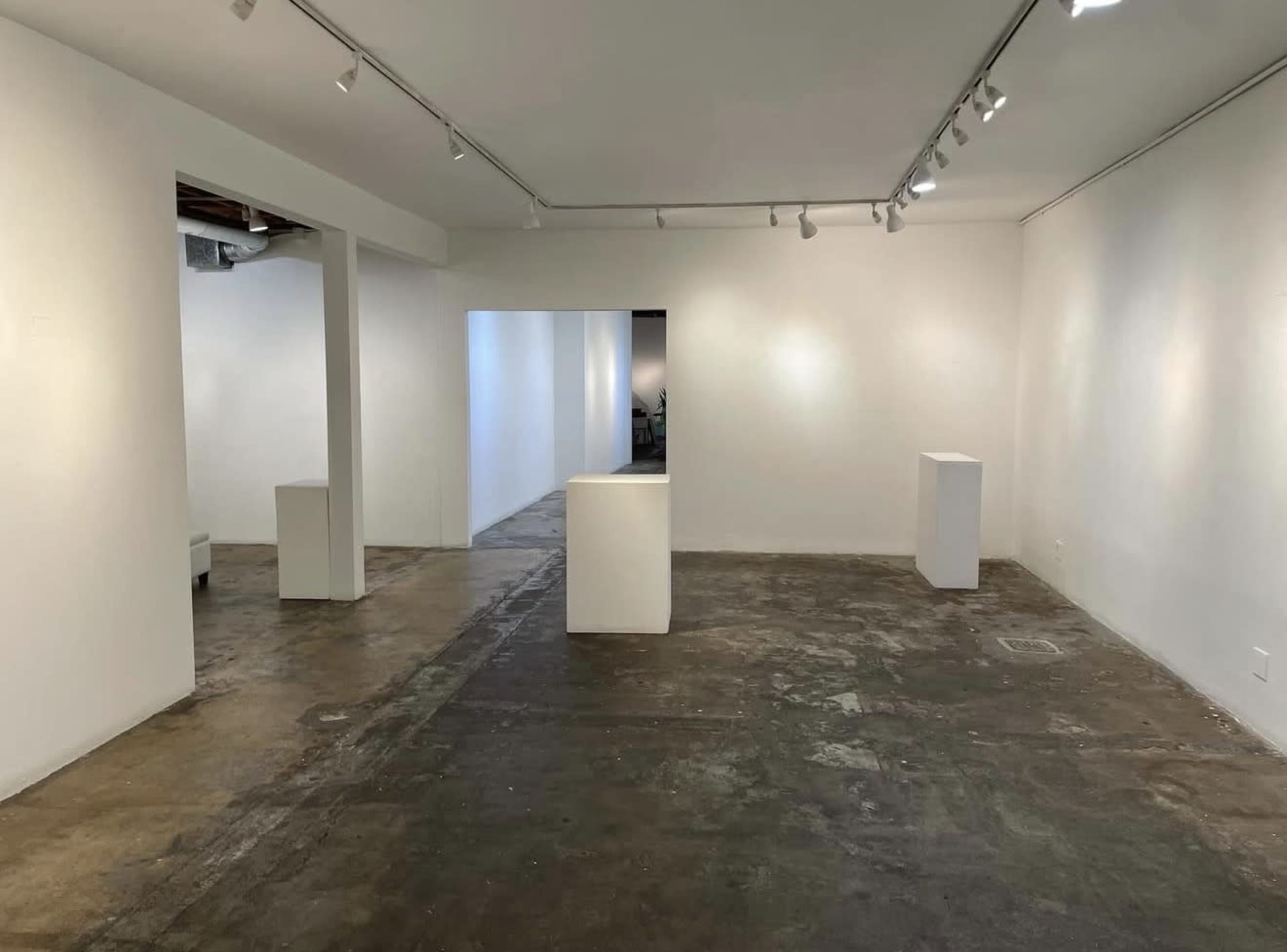 The image shows an empty art gallery with white walls and several white display pedestals on a concrete floor.