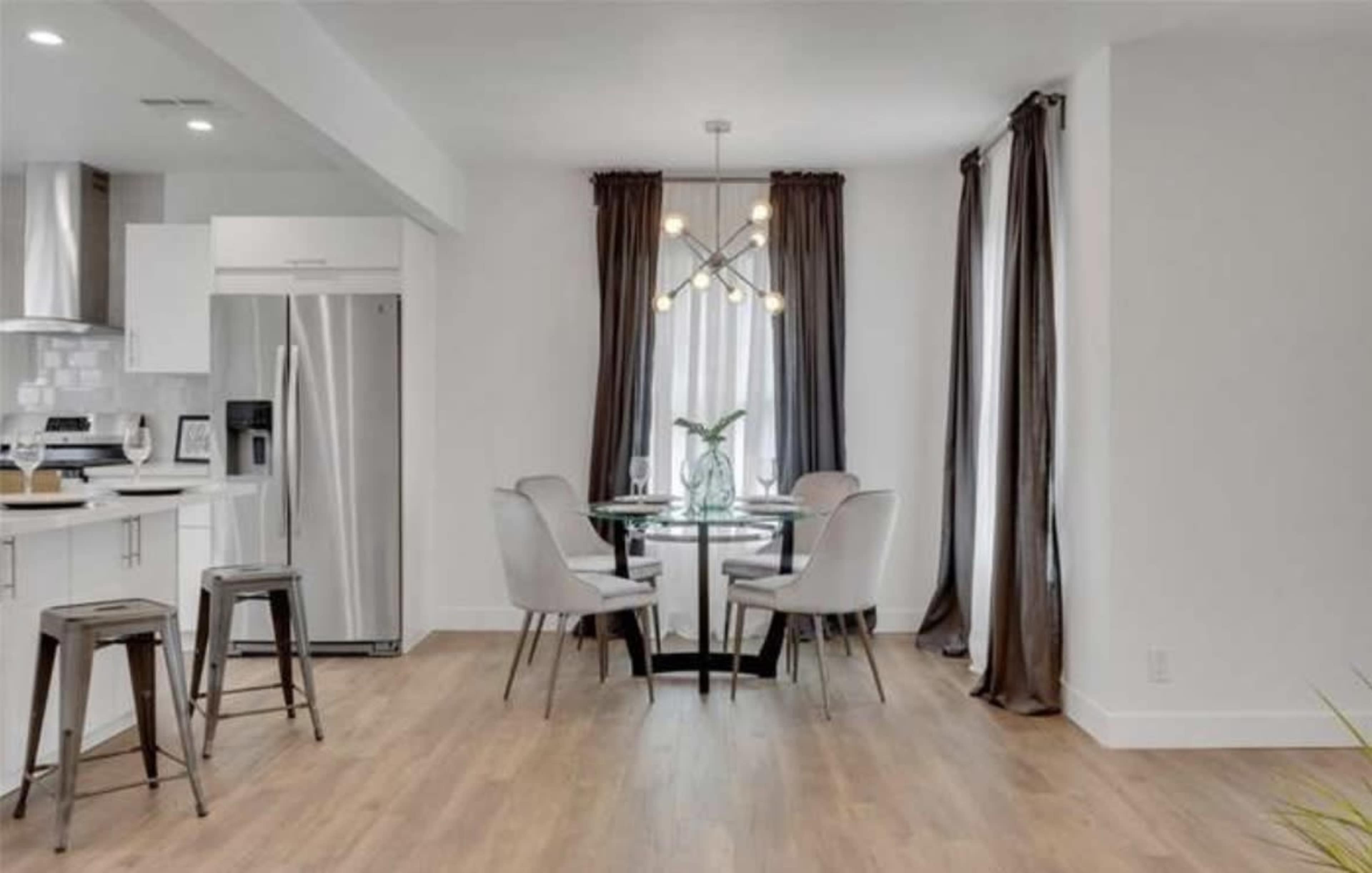 A modern dining area features a circular glass table surrounded by four upholstered chairs, with a pendant light overhead and large windows dressed in dark curtains.
