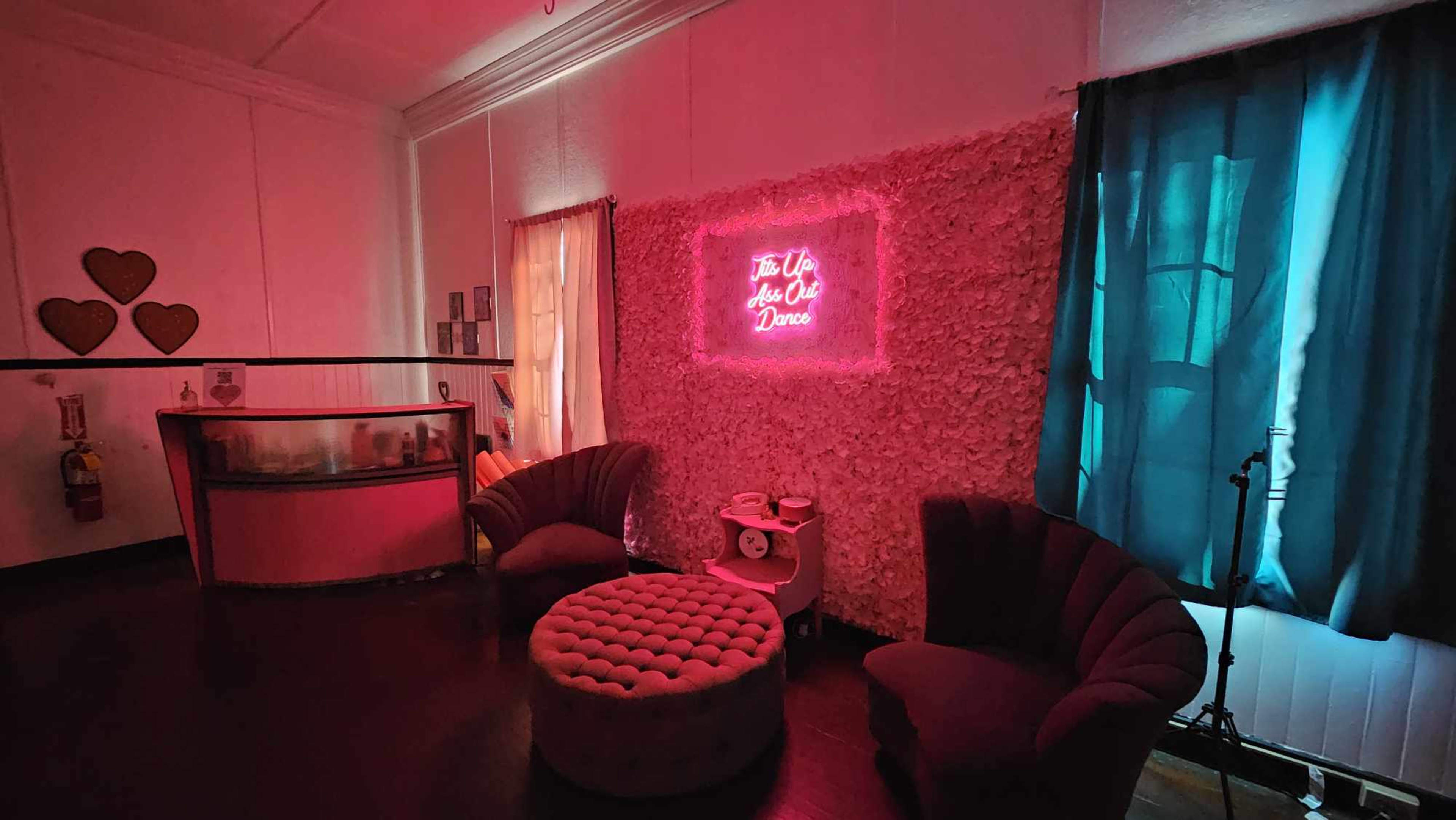 The image shows a cozy room with a pink neon sign on a textured wall, plush seating, and blue curtains.