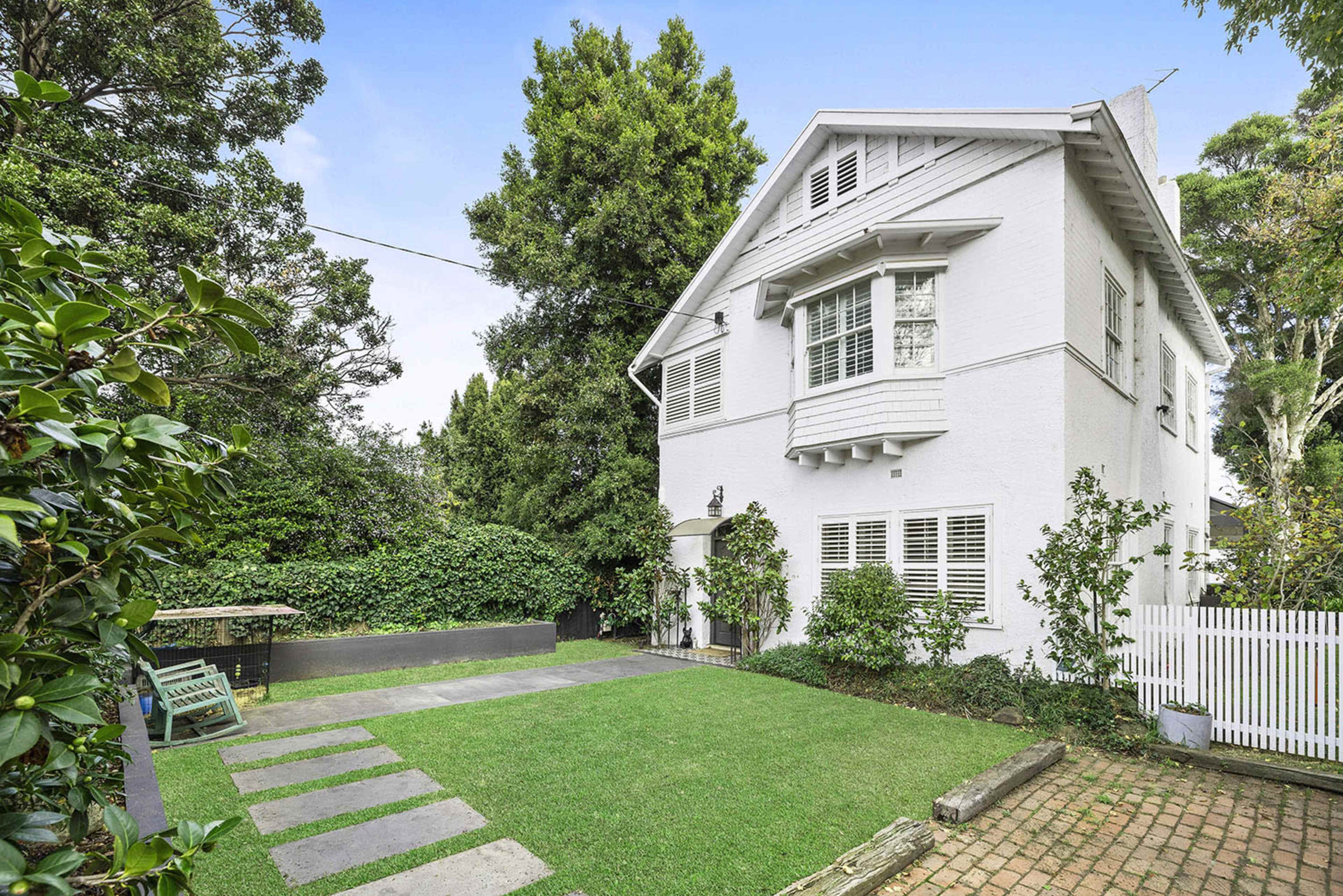 A white two-story house with multiple windows is surrounded by greenery and features a stone pathway leading to a lawn area.