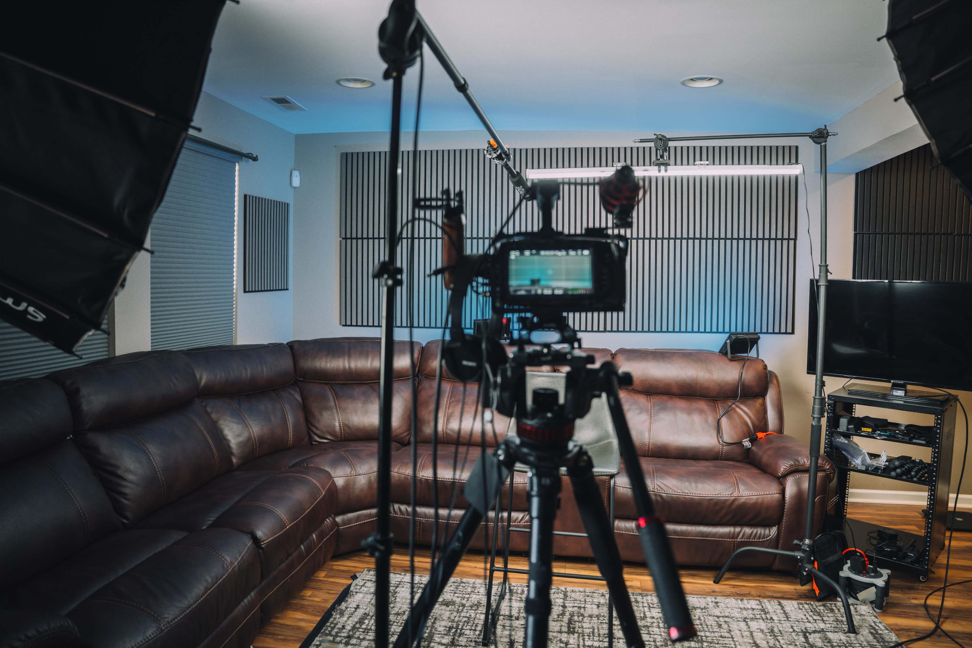 A camera on a tripod is positioned in a well-lit living room featuring a large brown sectional couch and various studio lights.