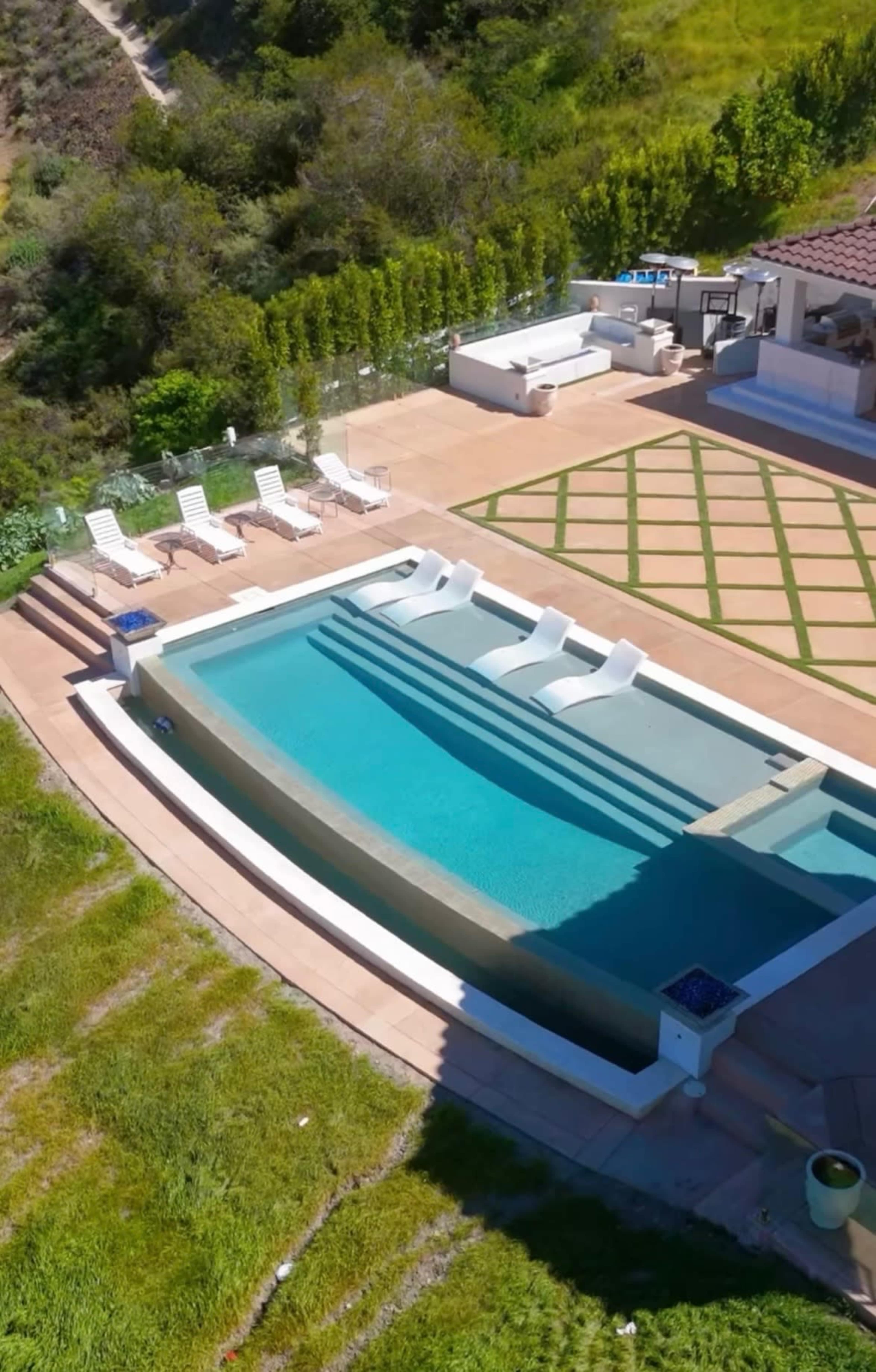 The image shows a modern outdoor space featuring a rectangular swimming pool with built-in steps, surrounded by lounge chairs and landscaped greenery.
