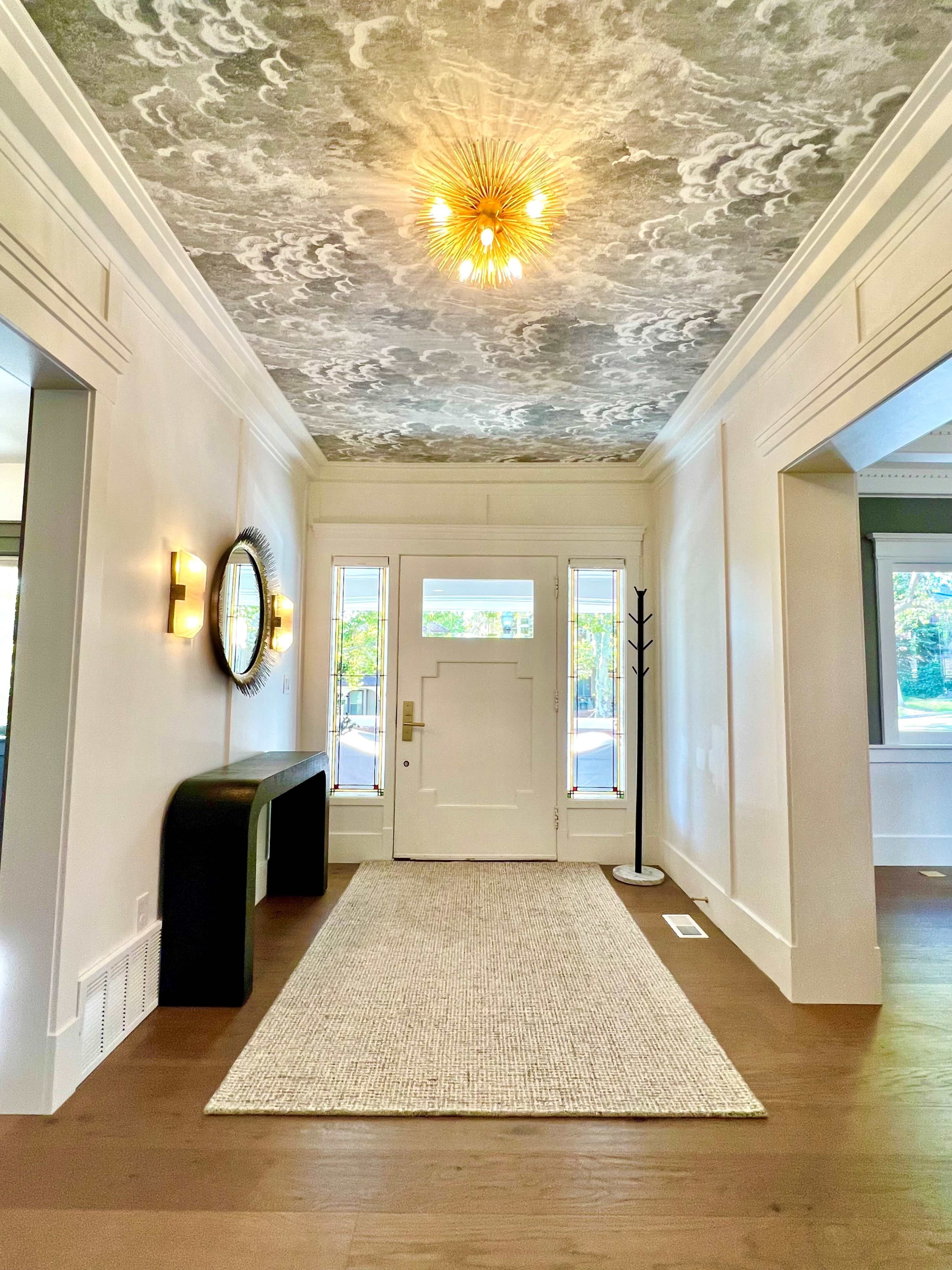 A well-lit hallway with a patterned ceiling, a round mirror on the wall, and a textured area rug leading to a front door.
